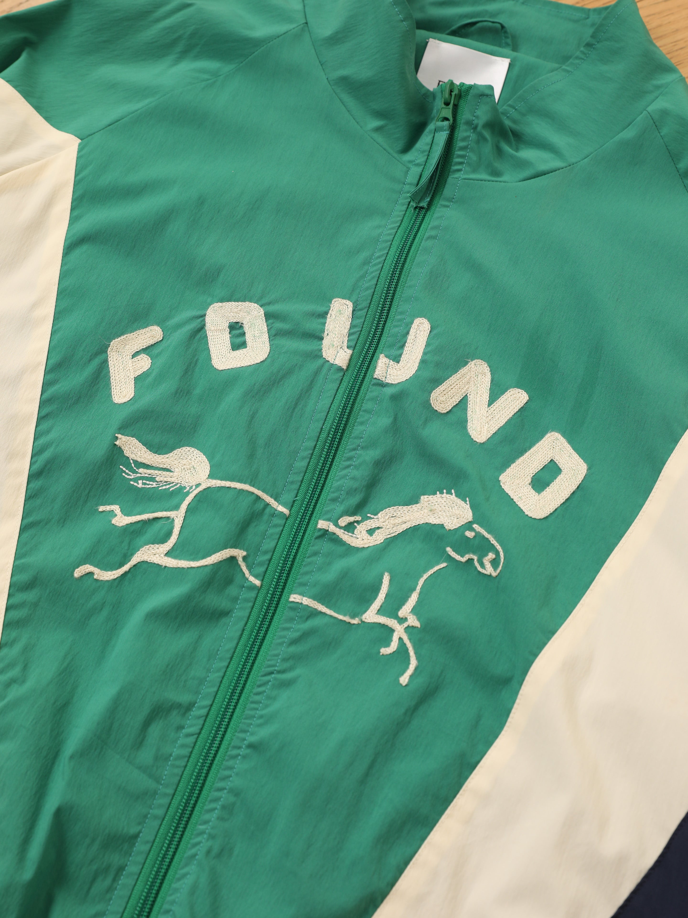The FOUND Crestline Sport Zip Jacket features green and white color blocking, a front zipper, and an embroidered horse with "FOUND" on the chest—a vintage-inspired lightweight windbreaker that blends comfort and style.
