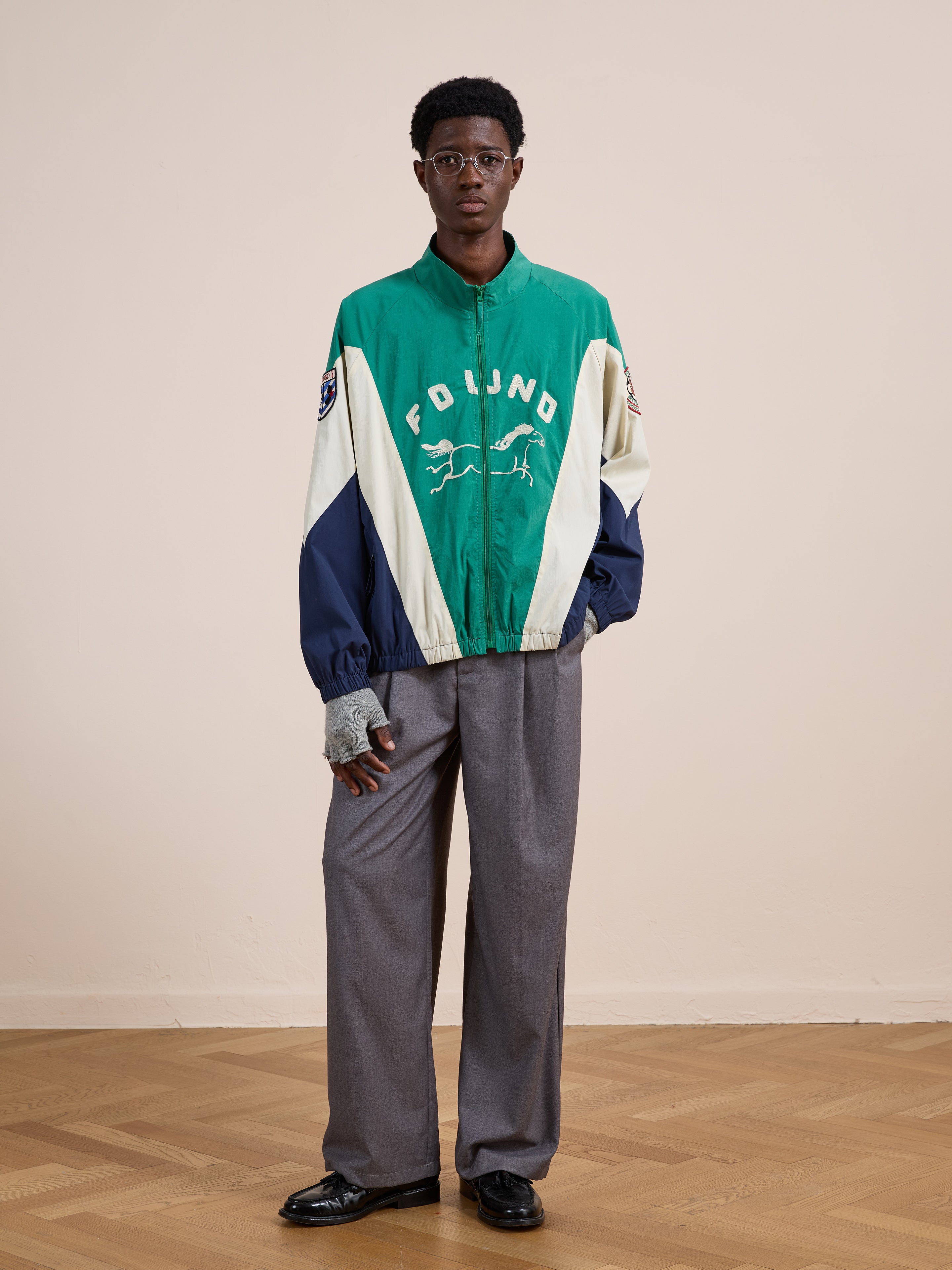 Indoors on a wooden floor, someone wearing glasses models the FOUND Crestline Sport Zip Jacket in green and white with graphic, paired with gray trousers and black shoes, standing against a plain light wall.