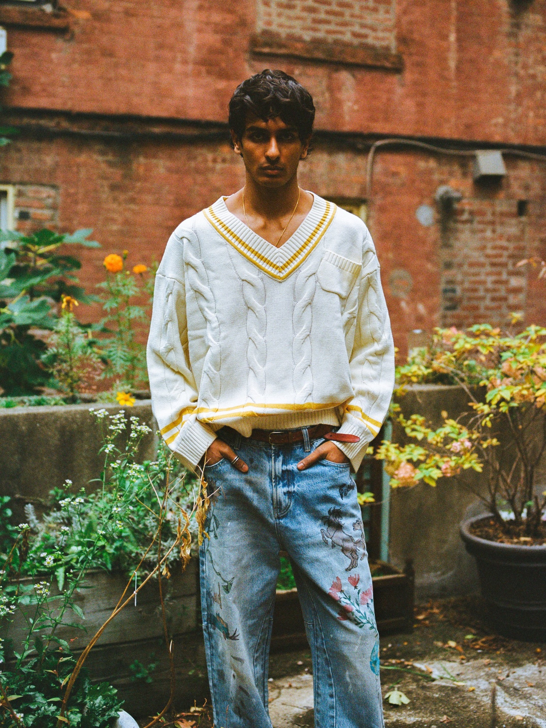 A man stands in an outdoor garden wearing the FOUND V-Neck Cricket Sweater with yellow trim and light blue floral jeans, with a brick wall and potted plants adding background charm.