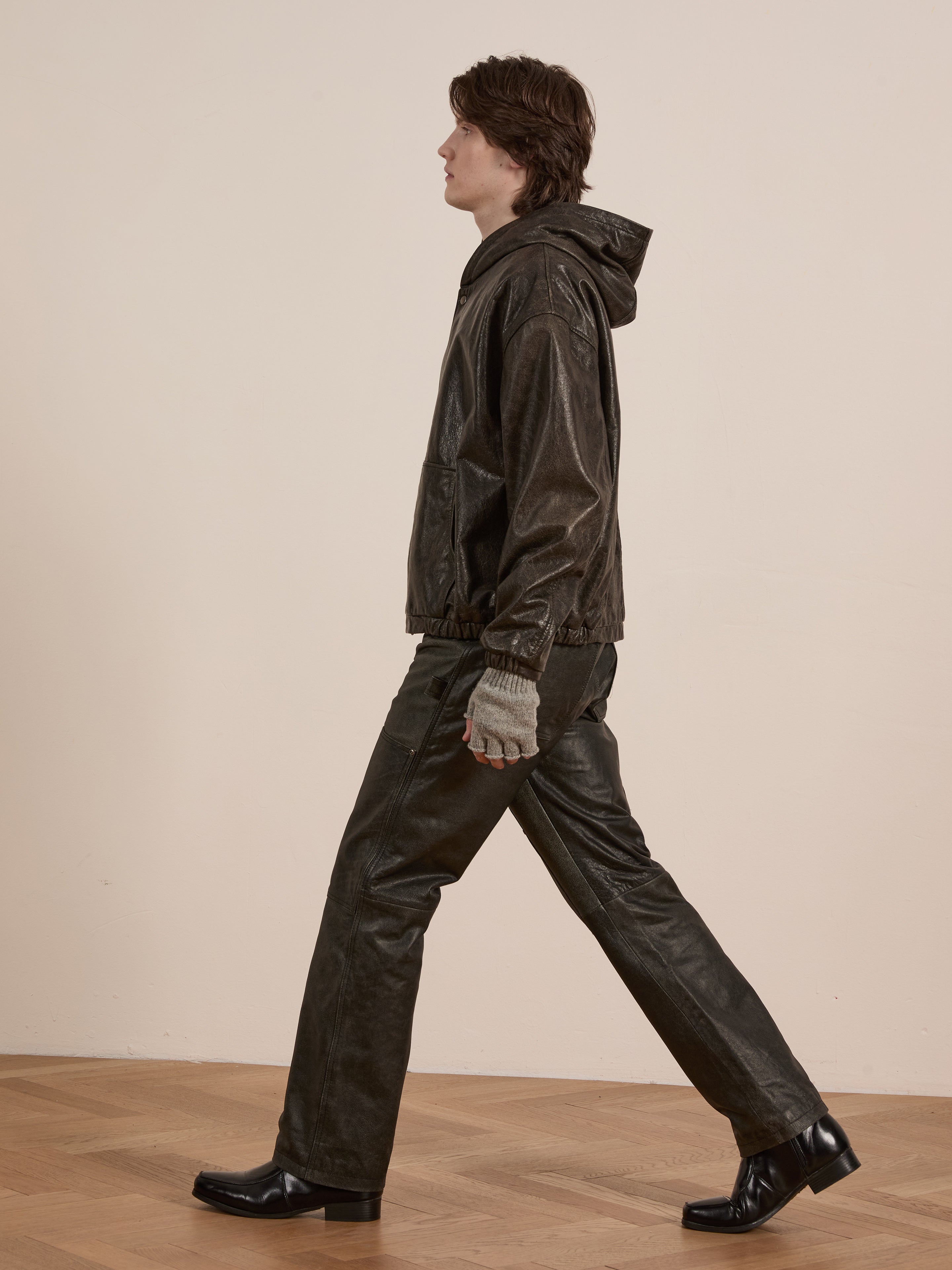 A person walks in profile wearing Found's Distressed Genuine Leather Hoodie with matching leather pants, gray gloves, and black shoes on a wooden floor against a plain background.