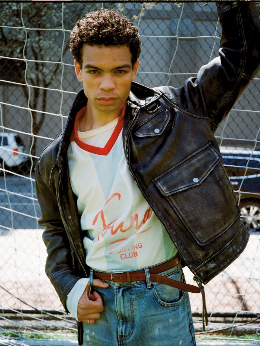 A young man stands before a soccer net, wearing Found's Distressed Leather Pocket Jacket over a white shirt with red trim and blue jeans, with cars and trees in the background.