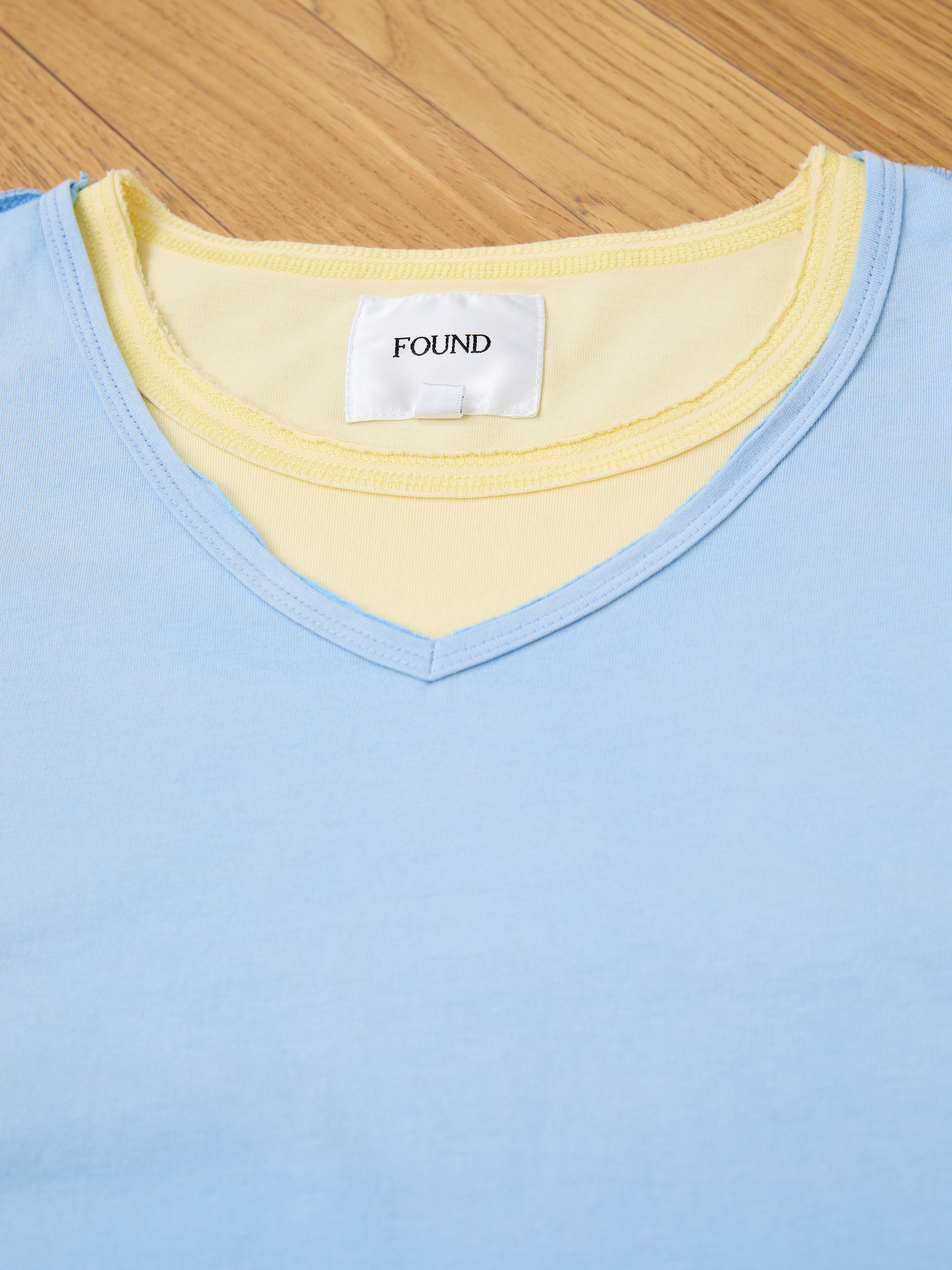 A light blue Double Layer LS Tee by FOUND with a sunfade wash is layered over a pale yellow unisex long sleeve tee, both lying flat on a wooden floor. The visible tag on the yellow tee reads "FOUND.