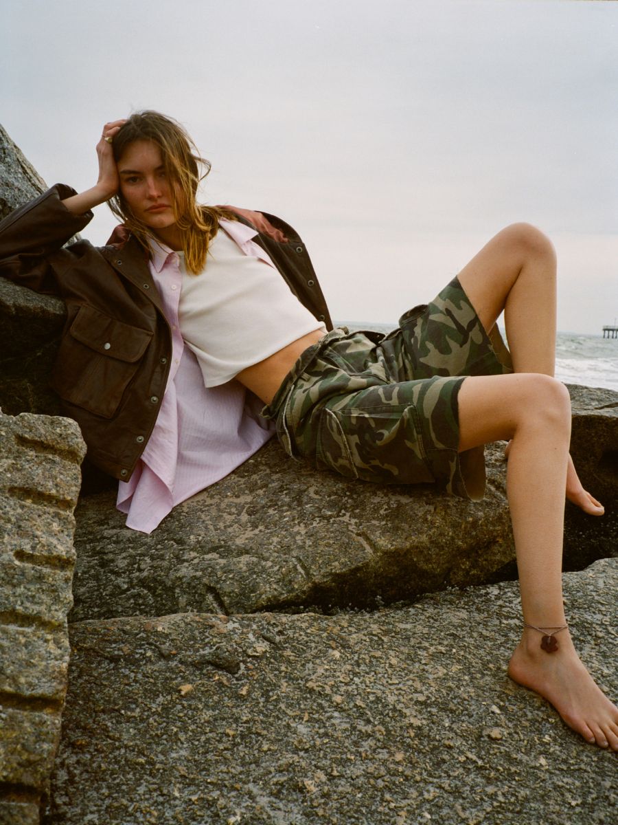 A person lounges on rocks by the shore, wearing a FOUND Dusk Distressed Genuine Leather Hoodie, a white crop top, pink shirt, camouflage shorts, and an anklet with bare feet.