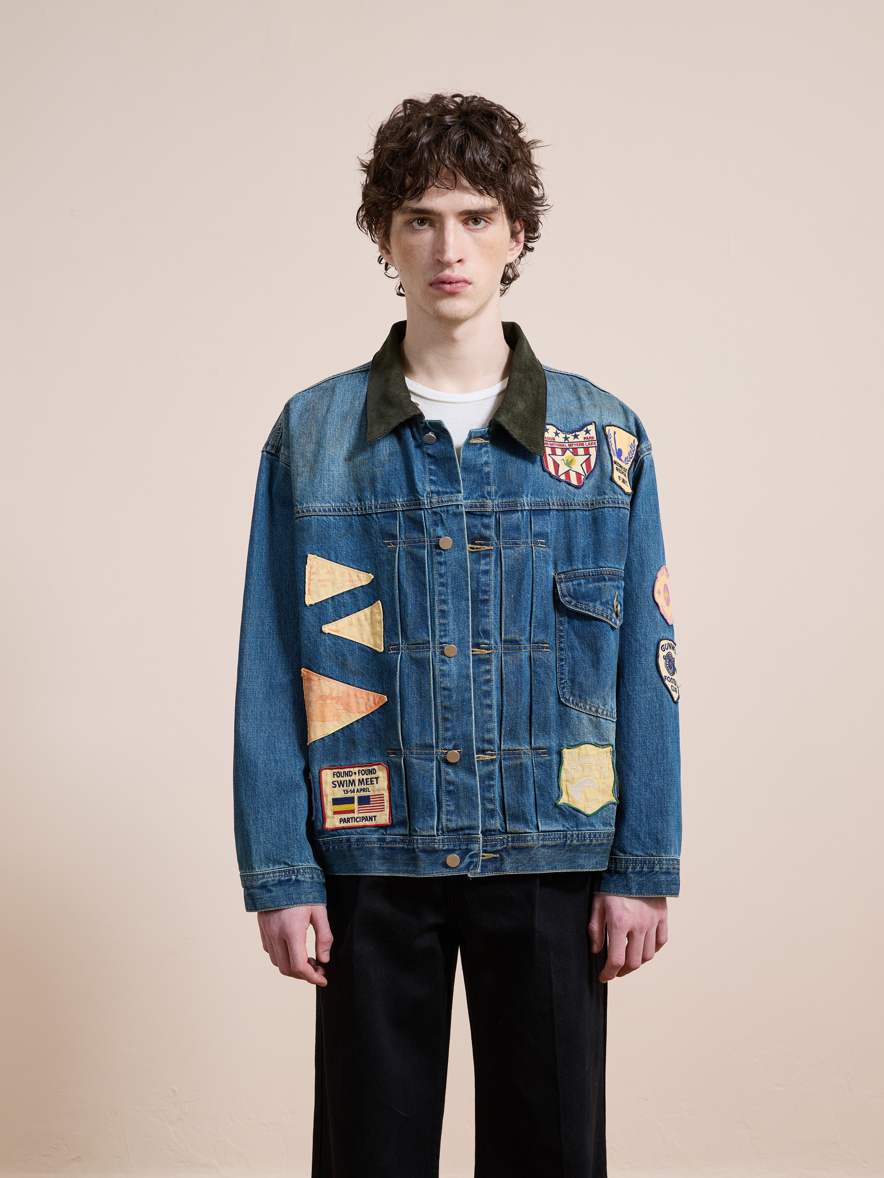 A person with short curly hair wears the FOUND Denim Patchwork Jacket and black pants, standing against a plain beige background.