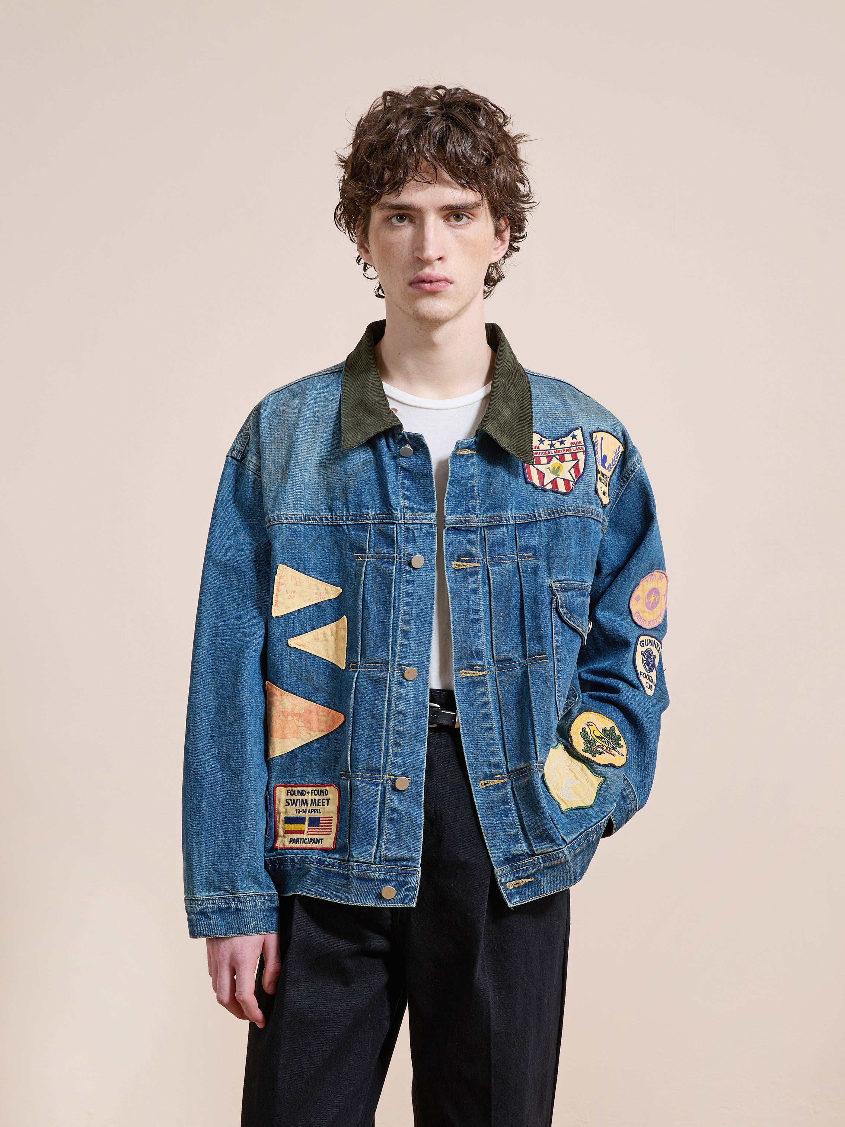 A young person with curly hair wears the FOUND Denim Patchwork Jacket over a white shirt and black pants, standing against a plain beige background.