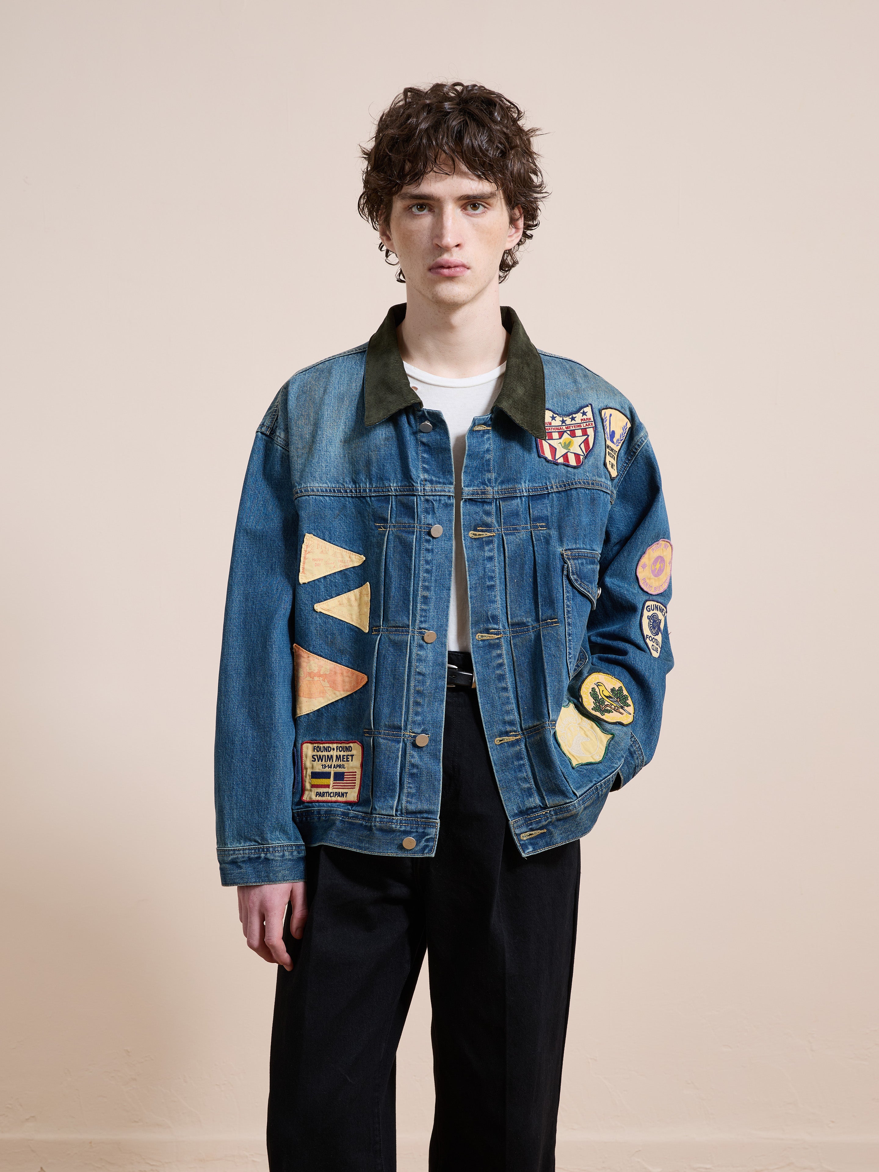 A person stands against a plain background wearing the FOUND Denim Patchwork Jacket, featuring vintage washed denim and colorful patches, paired with a white shirt and black pants.