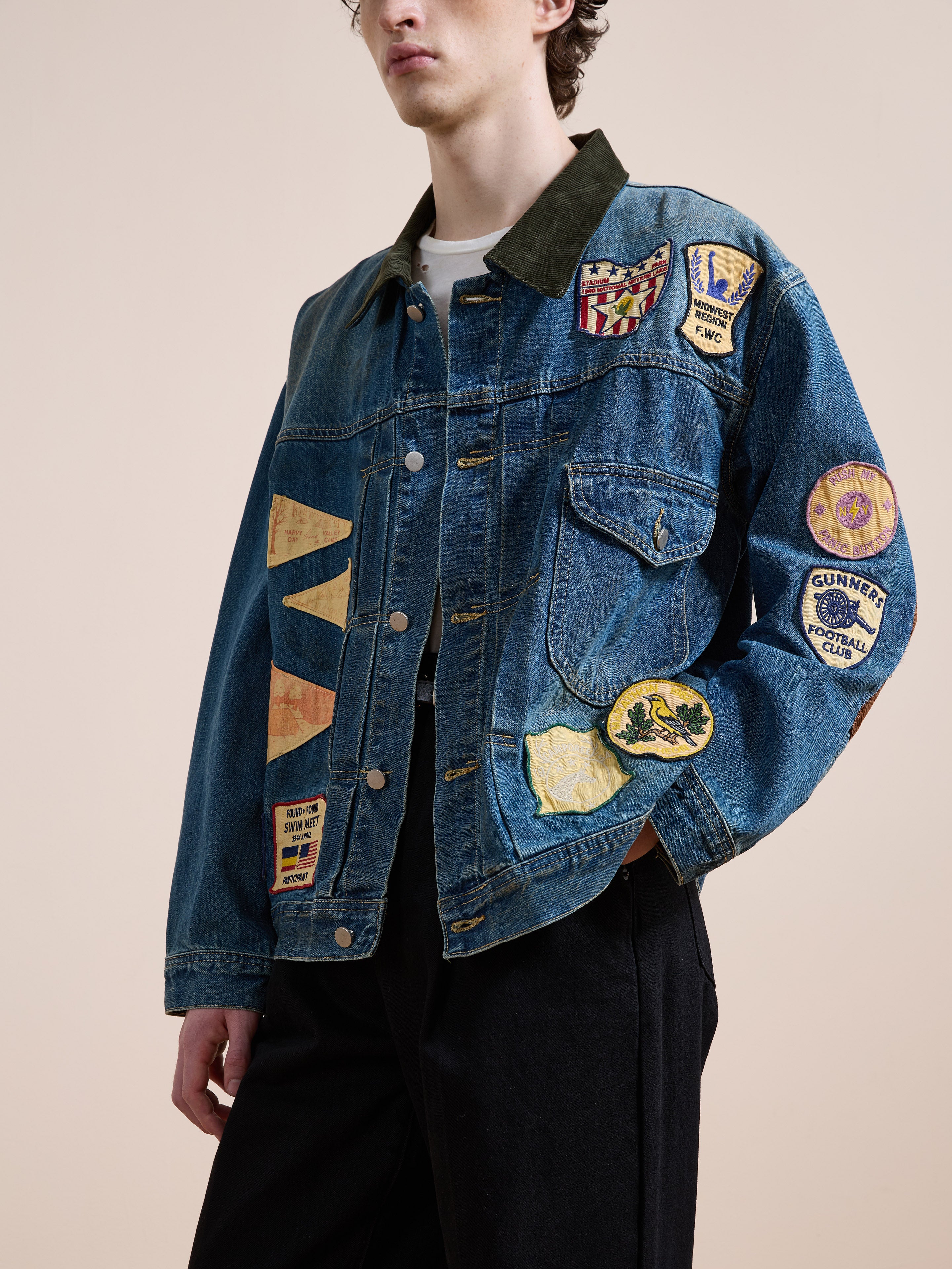 A person wearing the FOUND Denim Patchwork Jacket, featuring a vintage wash and colorful patches in a unisex relaxed fit, stands against a plain beige background.
