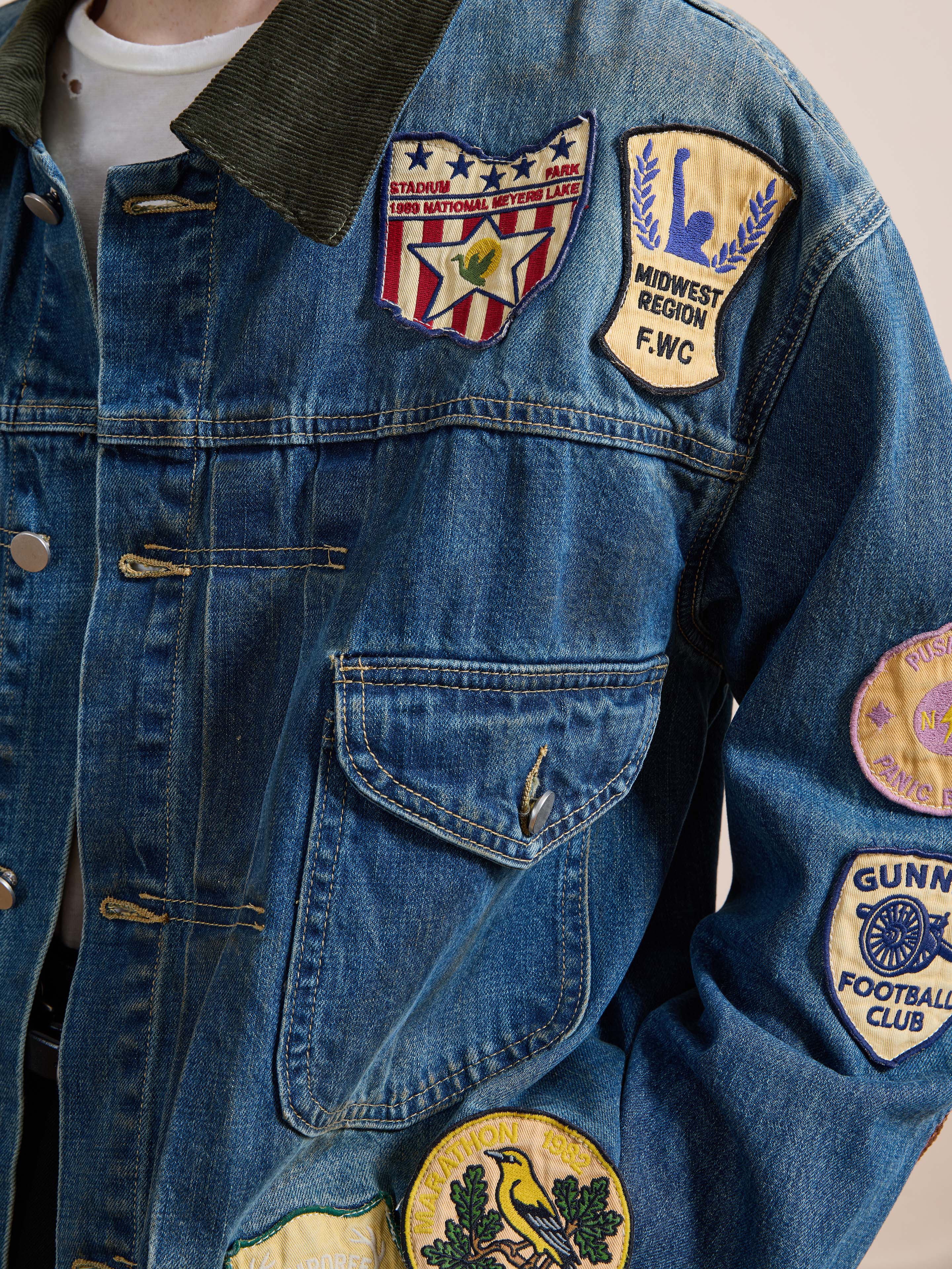 A person wears the FOUND Denim Patchwork Jacket, a vintage-washed denim piece featuring colorful embroidered patches and badges with text and emblem designs on the chest, pocket, and sleeve.