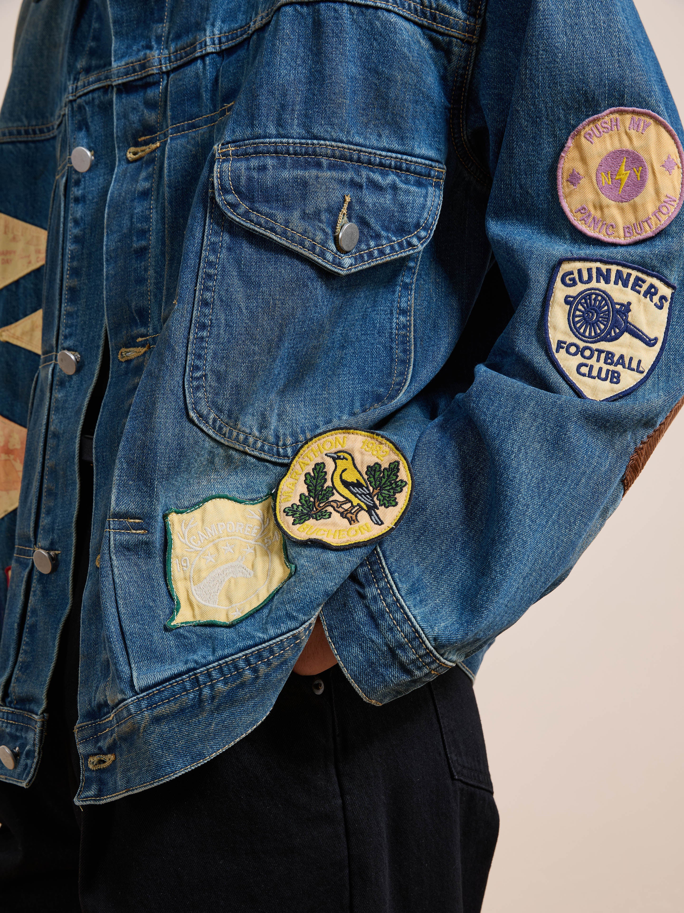 A person wears the FOUND Denim Patchwork Jacket, featuring vintage-washed denim adorned with colorful embroidered sports and nature-themed patches.