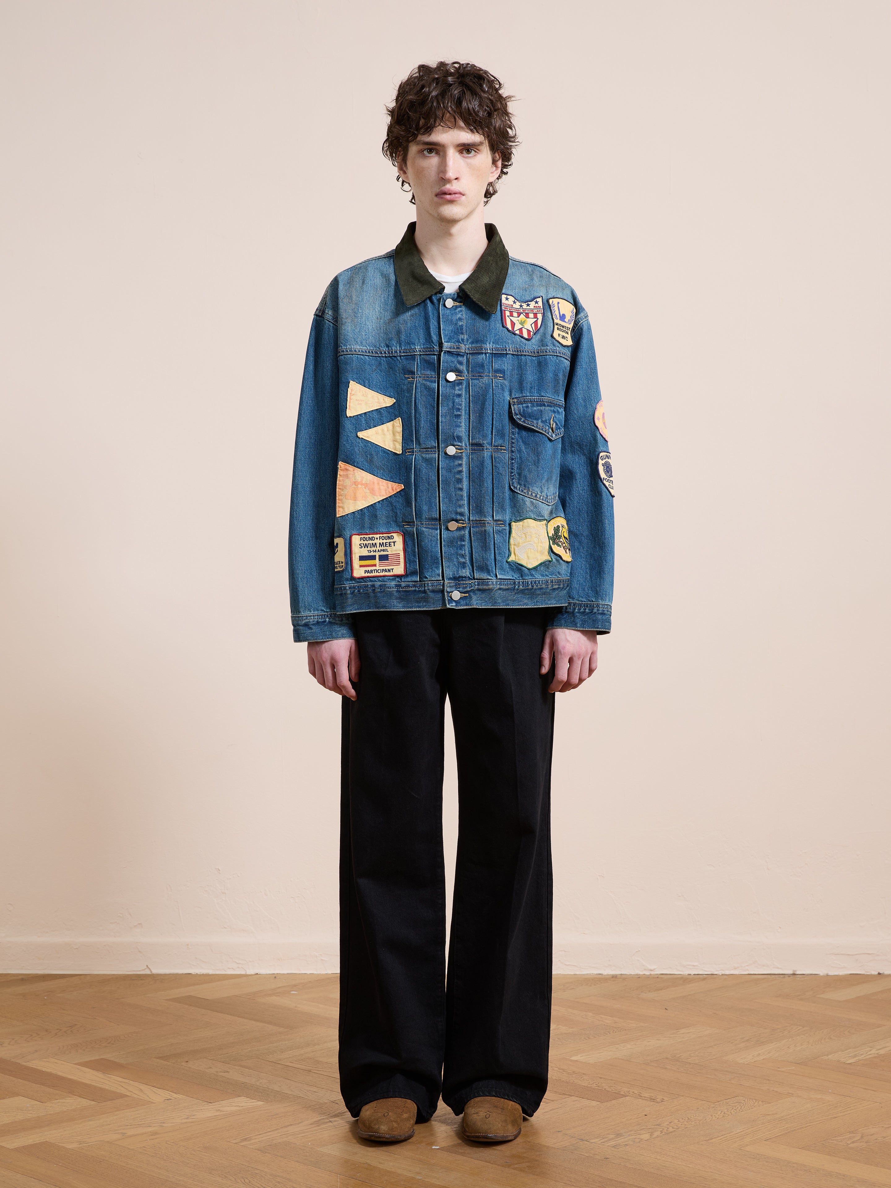 Indoors, a person models the FOUND Denim Patchwork Jacket with wide black pants and brown shoes, posed against a plain light wall and wooden floor.