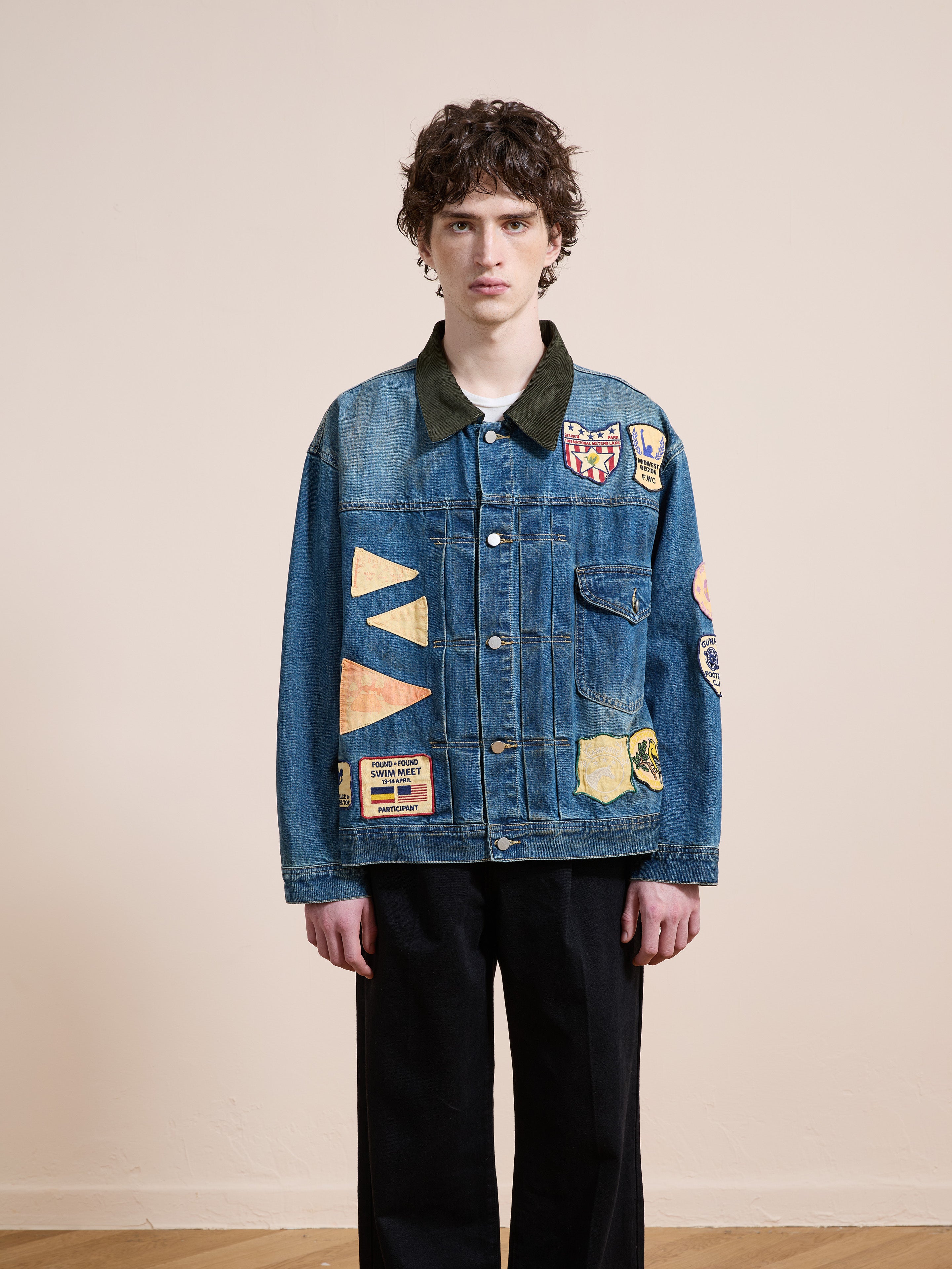 A person with short, curly hair wears the FOUND Denim Patchwork Jacket with colorful patches and black pants, standing against a plain beige wall.