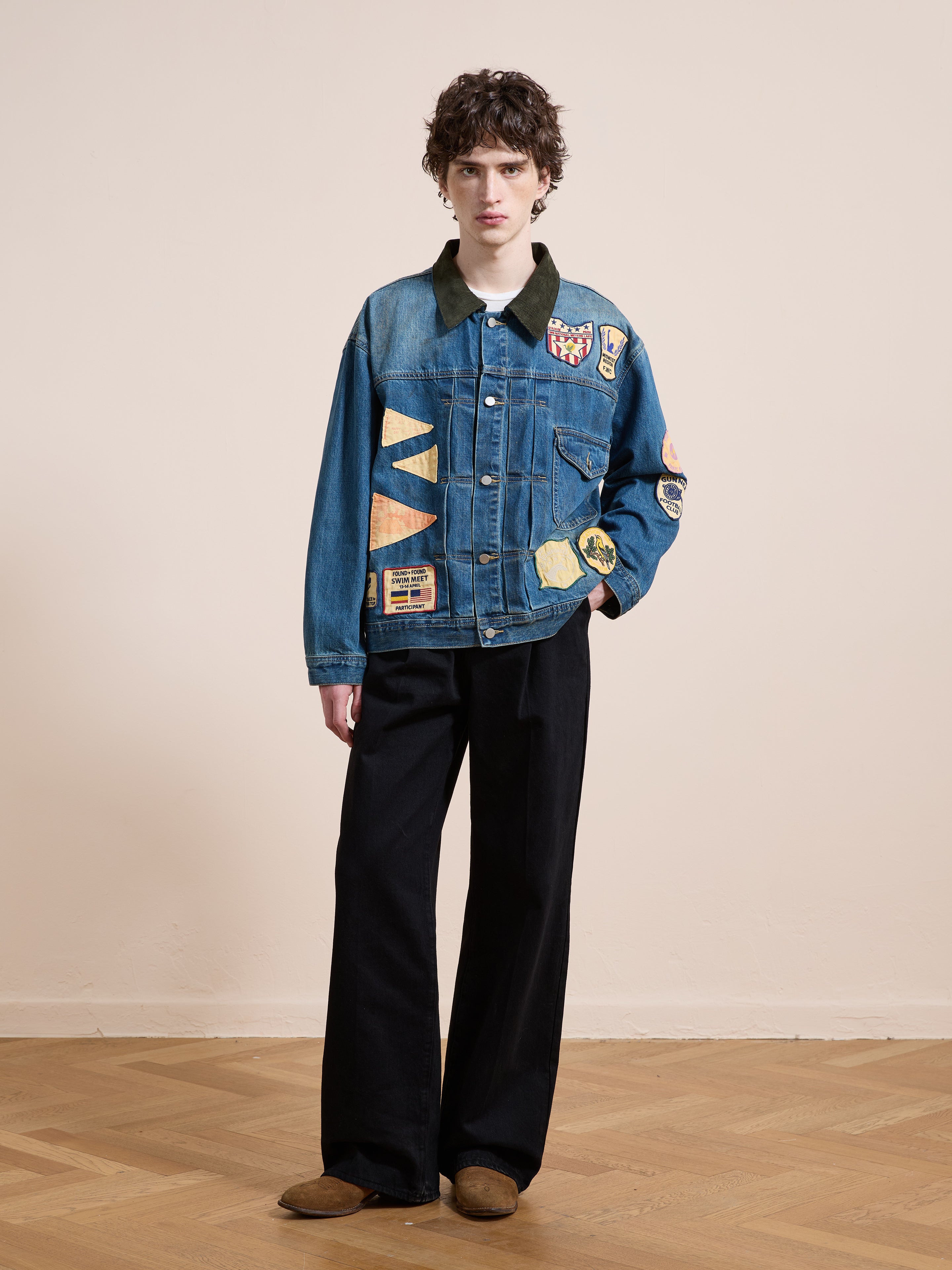 A person stands on a wooden floor against a light wall, wearing FOUND’s Denim Patchwork Jacket, paired with wide-leg black pants and brown shoes.