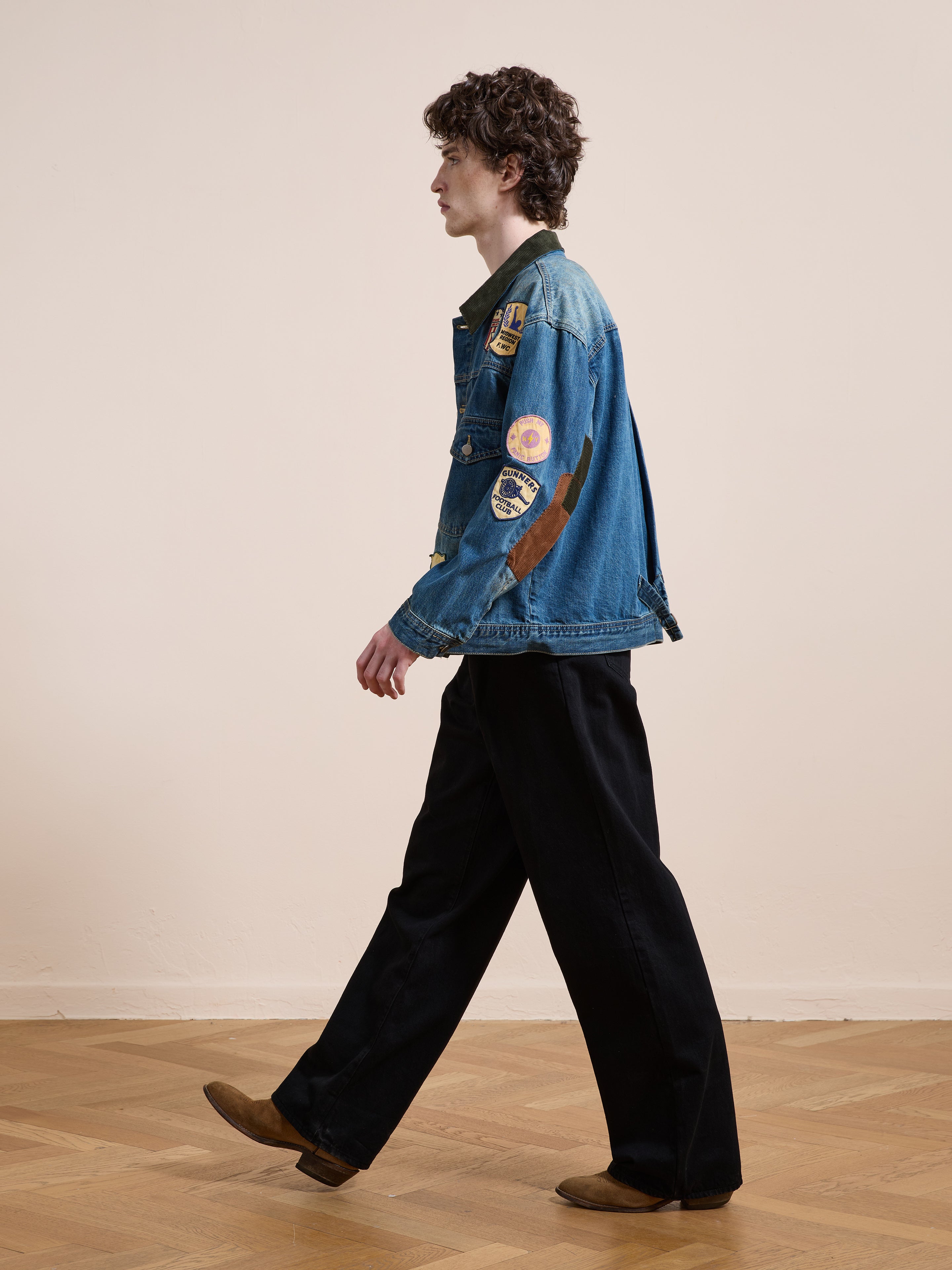A person with curly hair walks indoors, wearing the FOUND Denim Patchwork Jacket, black pants, and brown shoes.