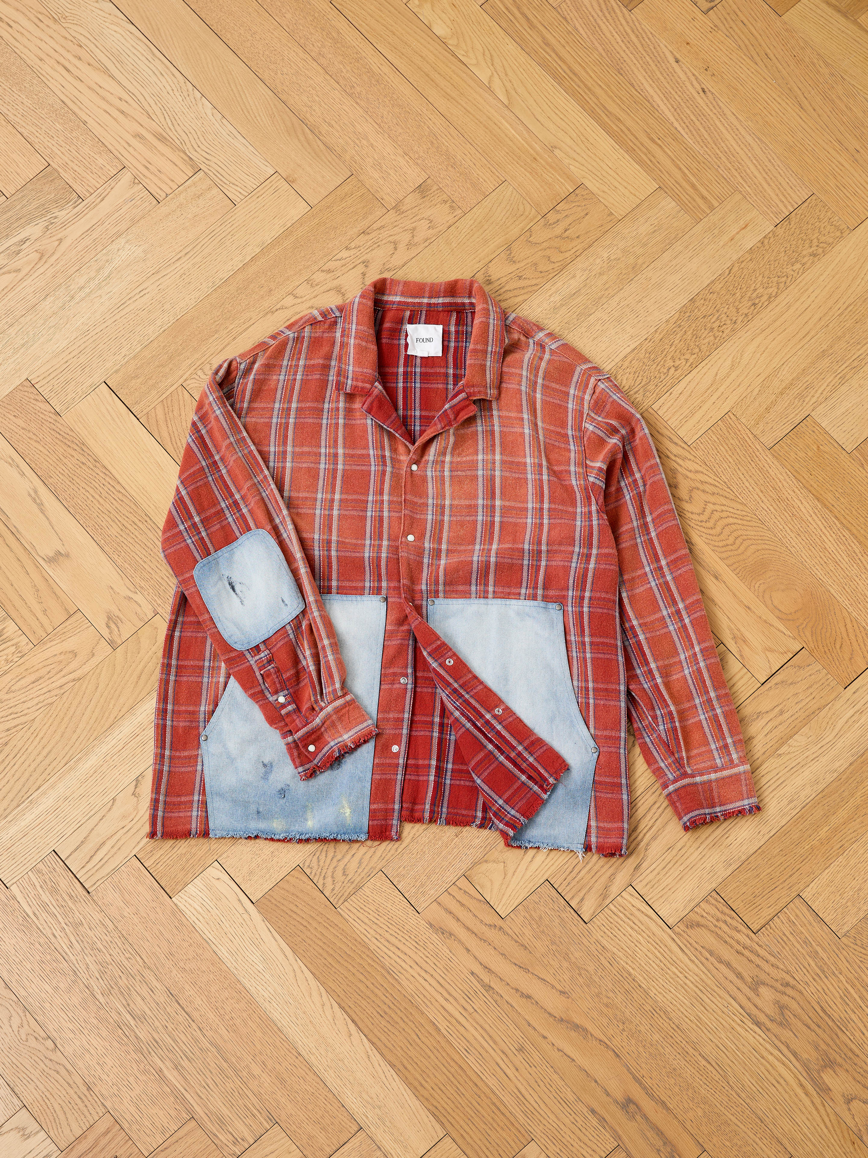 The FOUND Plaid Denim Shirt features red plaid fabric, denim carpenter pockets, and patchwork on the elbows and lower front, shown laid flat on a wooden herringbone floor—unisex workwear style.