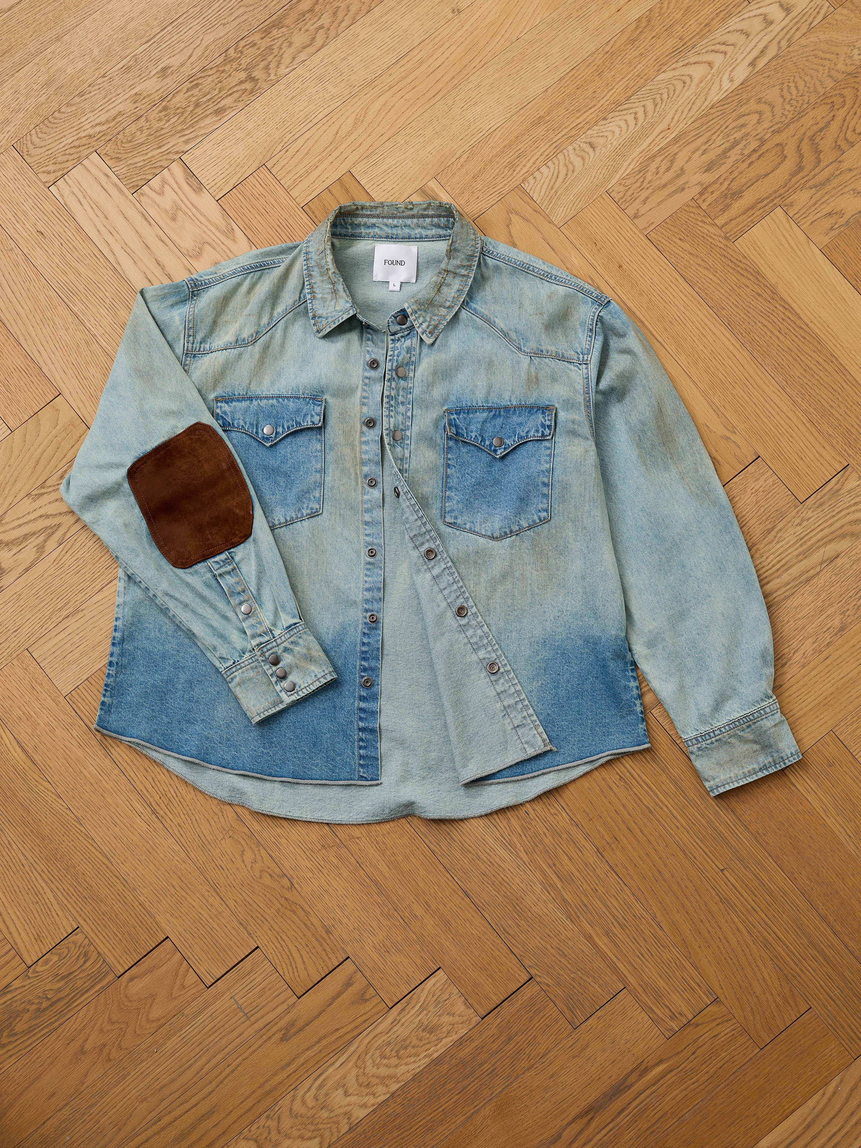 The FOUND Denim Western Snap Button Shirt in light blue features a unisex relaxed fit, snap closures, two chest pockets, and a brown suede elbow patch on the left sleeve, shown laid flat on a wooden parquet floor.