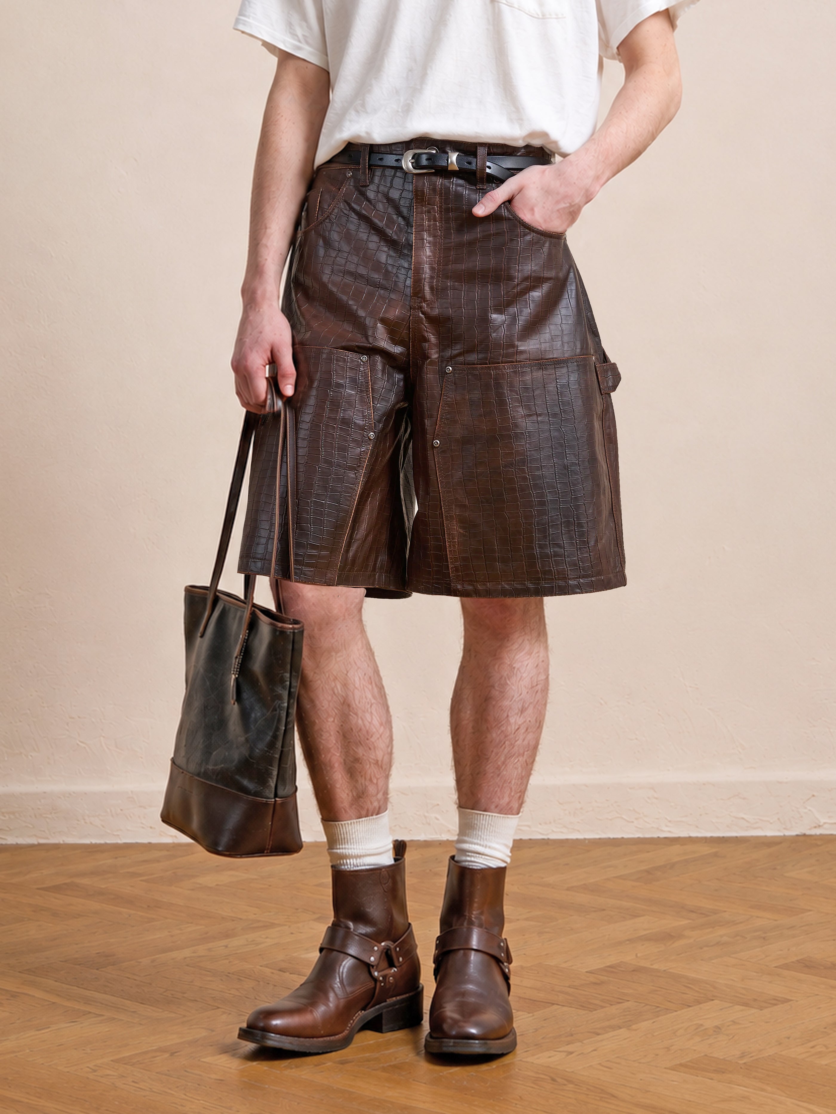 A person wearing a white t-shirt, FOUND Distressed Croc Embossed Leather Shorts, brown leather boots, and holding a brown leather tote bag stands on a wooden floor.