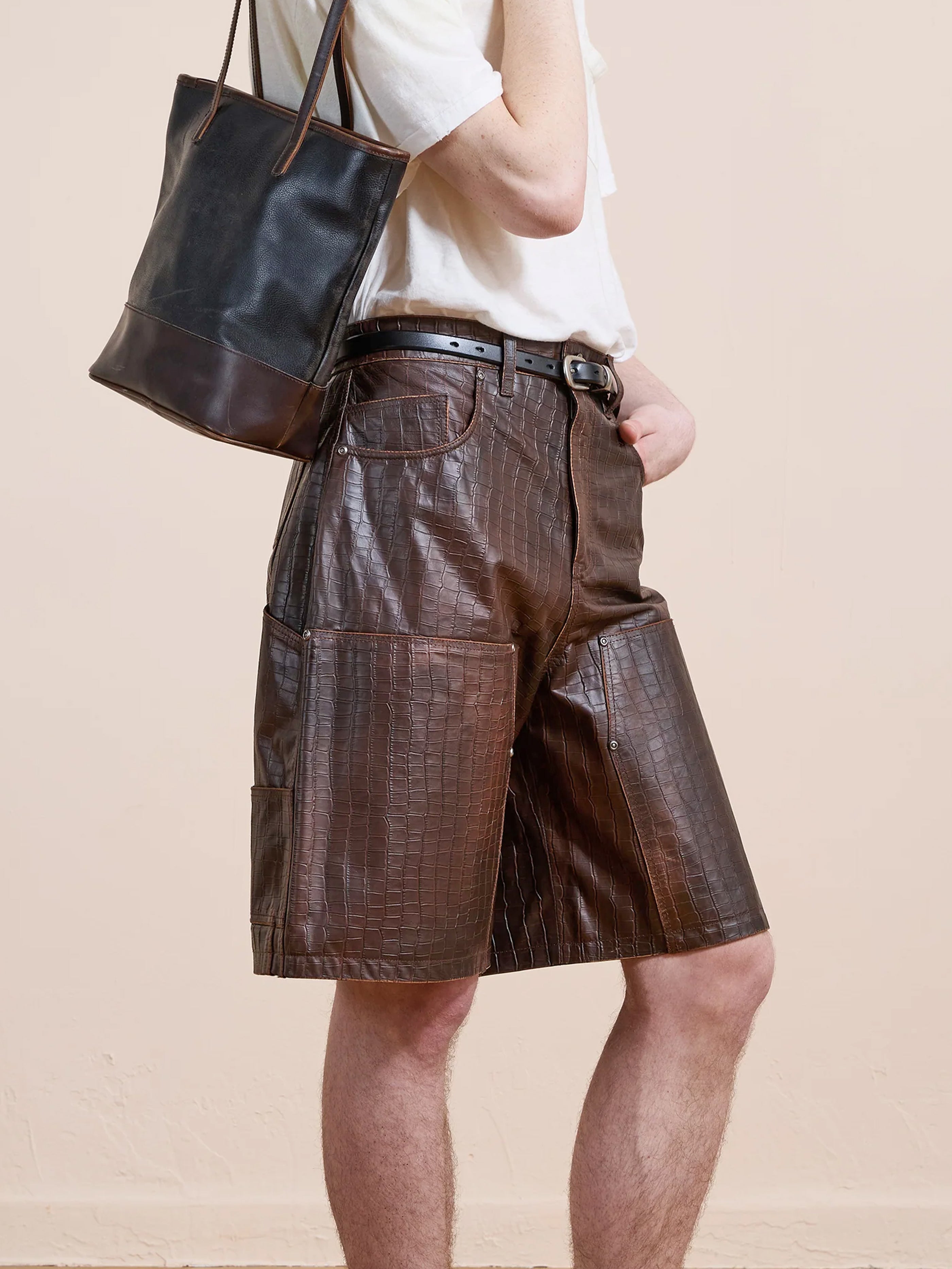 A person stands against a plain beige background wearing a white t-shirt, FOUND Distressed Croc Embossed Leather Shorts, and carrying a black and brown tote bag.