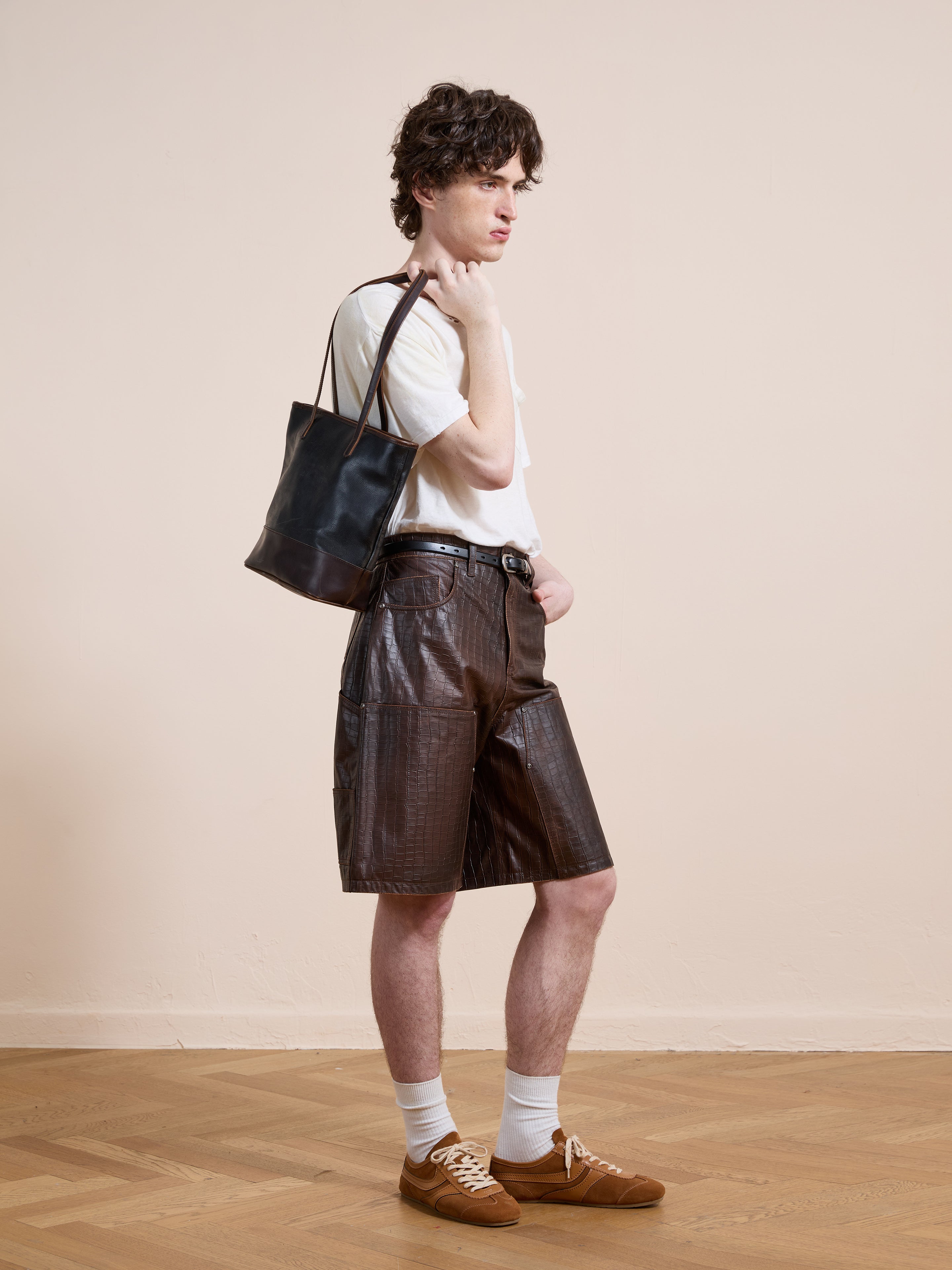 A person stands indoors wearing a white shirt, FOUND Distressed Croc Embossed Leather Shorts, white socks, brown sneakers, and carrying a black shoulder bag.