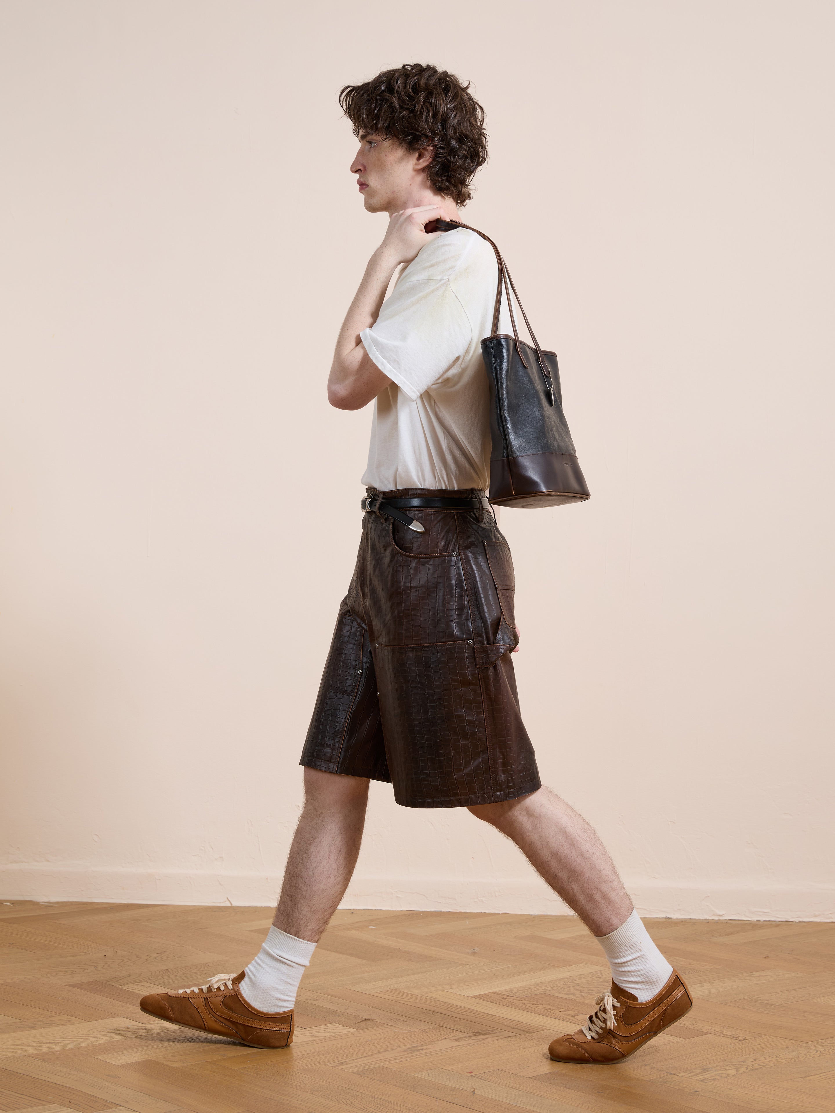 A person wearing a white t-shirt, FOUND Distressed Croc Embossed Leather Shorts, white socks, brown shoes, and carrying a black shoulder bag walks indoors.