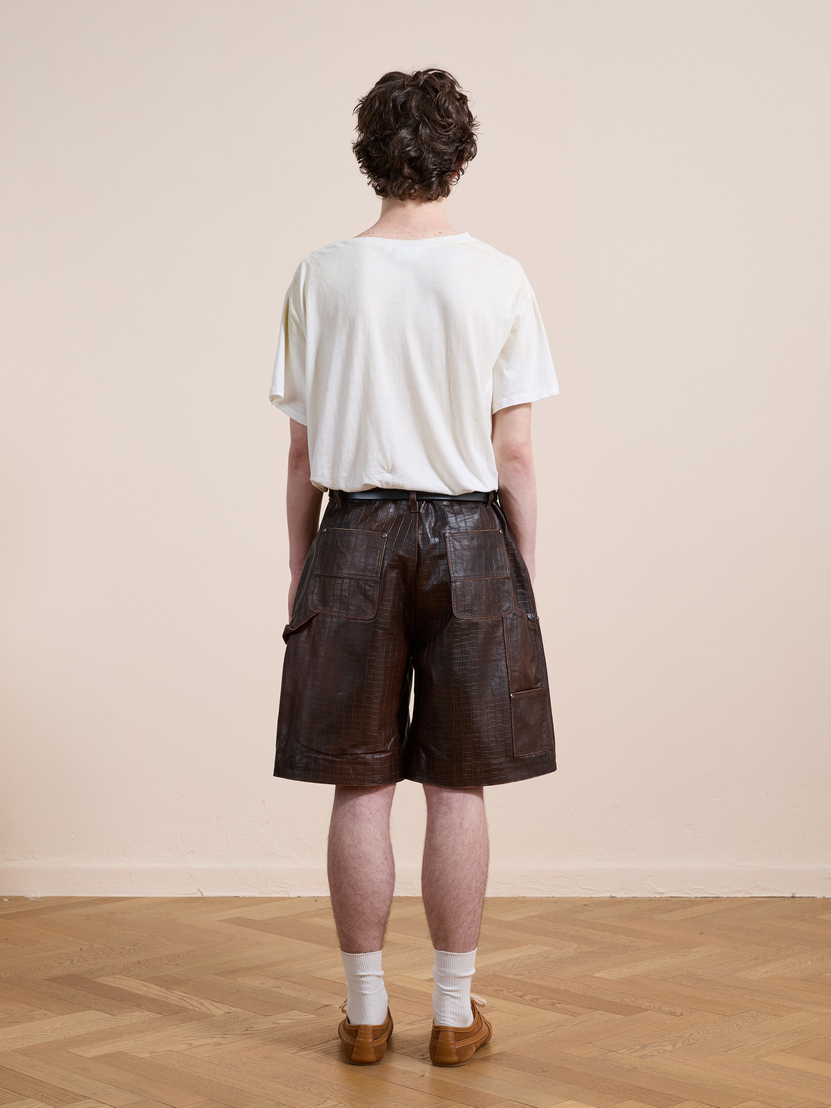 A person stands indoors on a wooden floor, facing away, wearing a white t-shirt, FOUND Distressed Croc Embossed Leather Shorts in dark brown, white socks, and tan shoes.