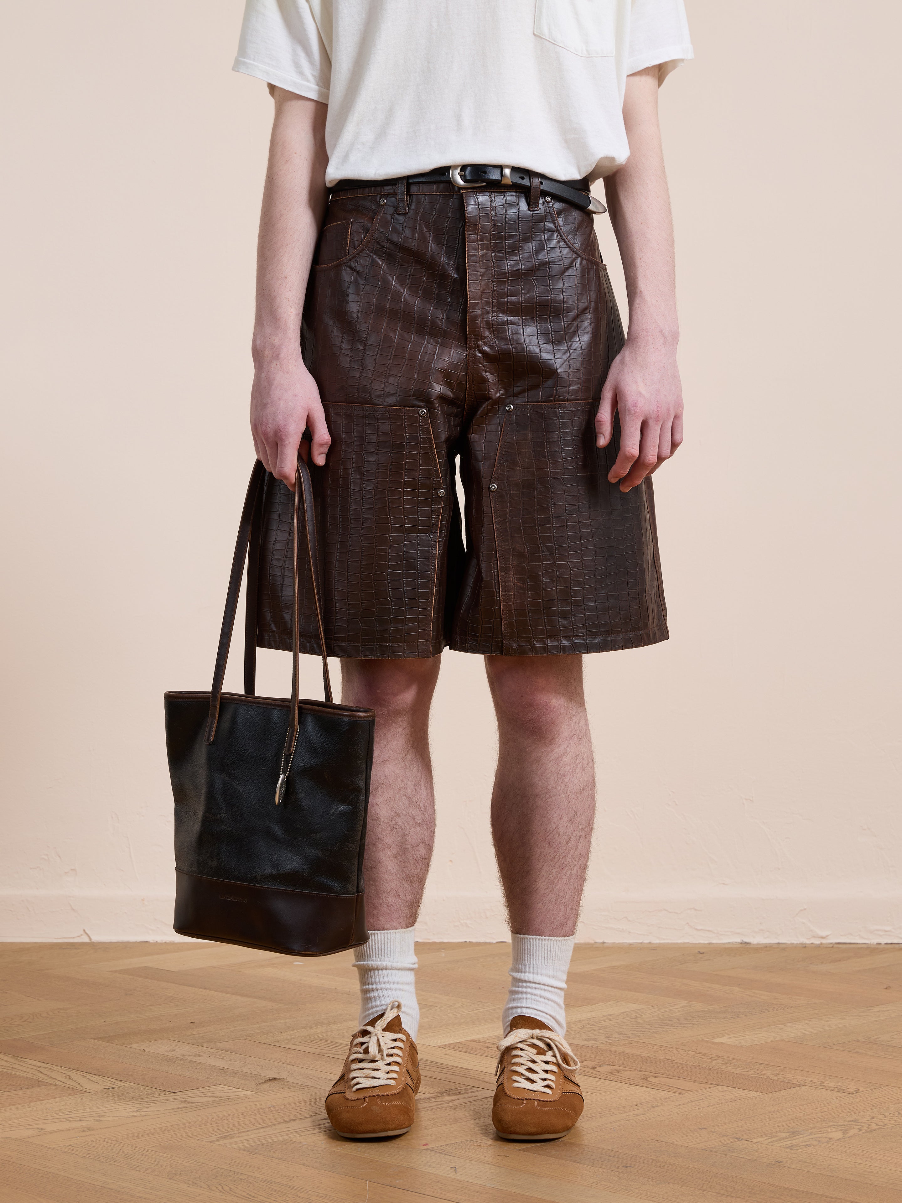 A person wears a white t-shirt, FOUND Distressed Croc Embossed Leather Shorts with a brown belt, white socks, brown sneakers, and holds a black leather tote bag while standing on a wooden floor against a beige wall.