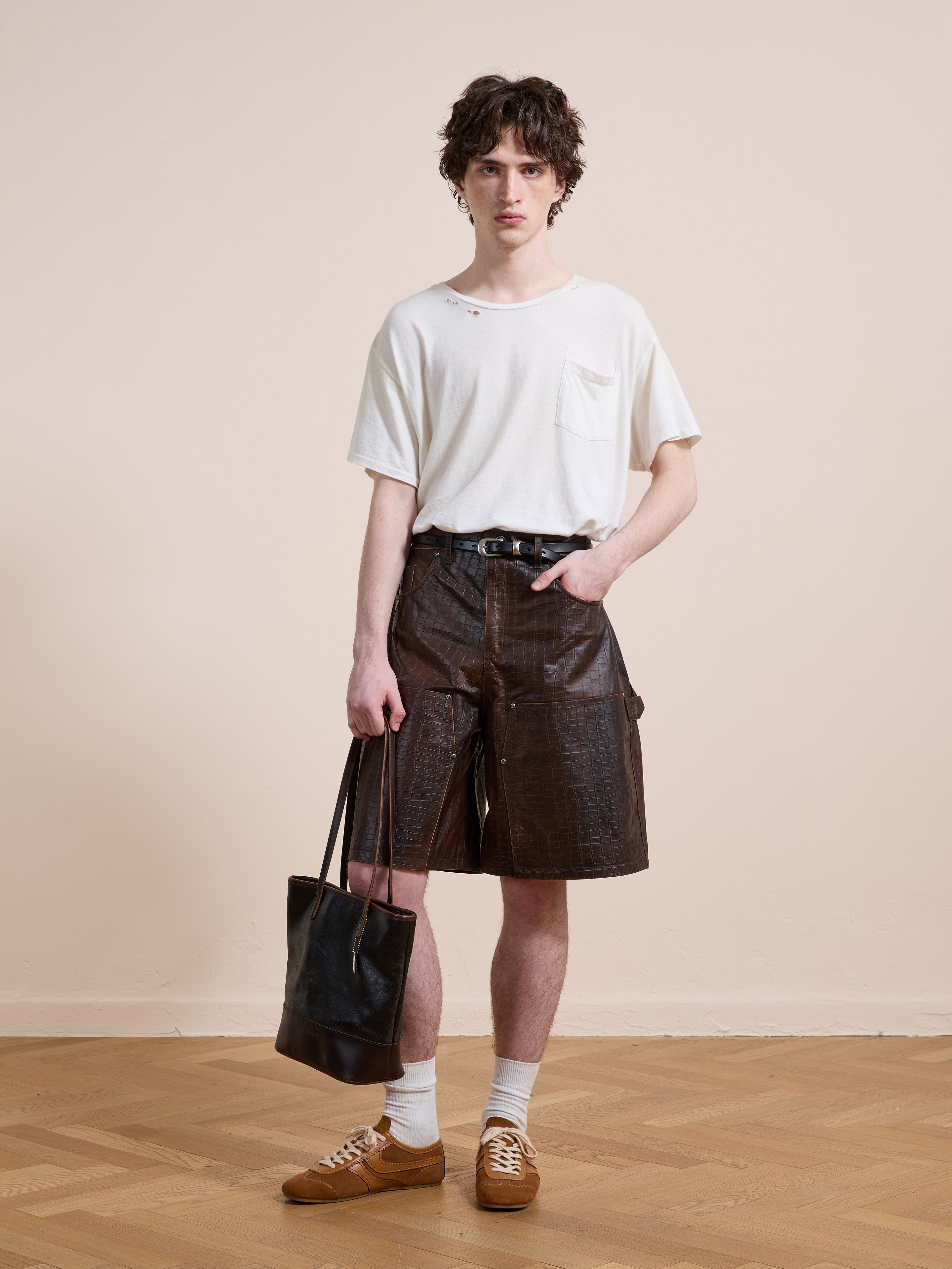 Indoors, a person wears a white t-shirt, FOUND Distressed Croc Embossed Leather Shorts, white socks, brown sneakers, and holds a black tote bag.