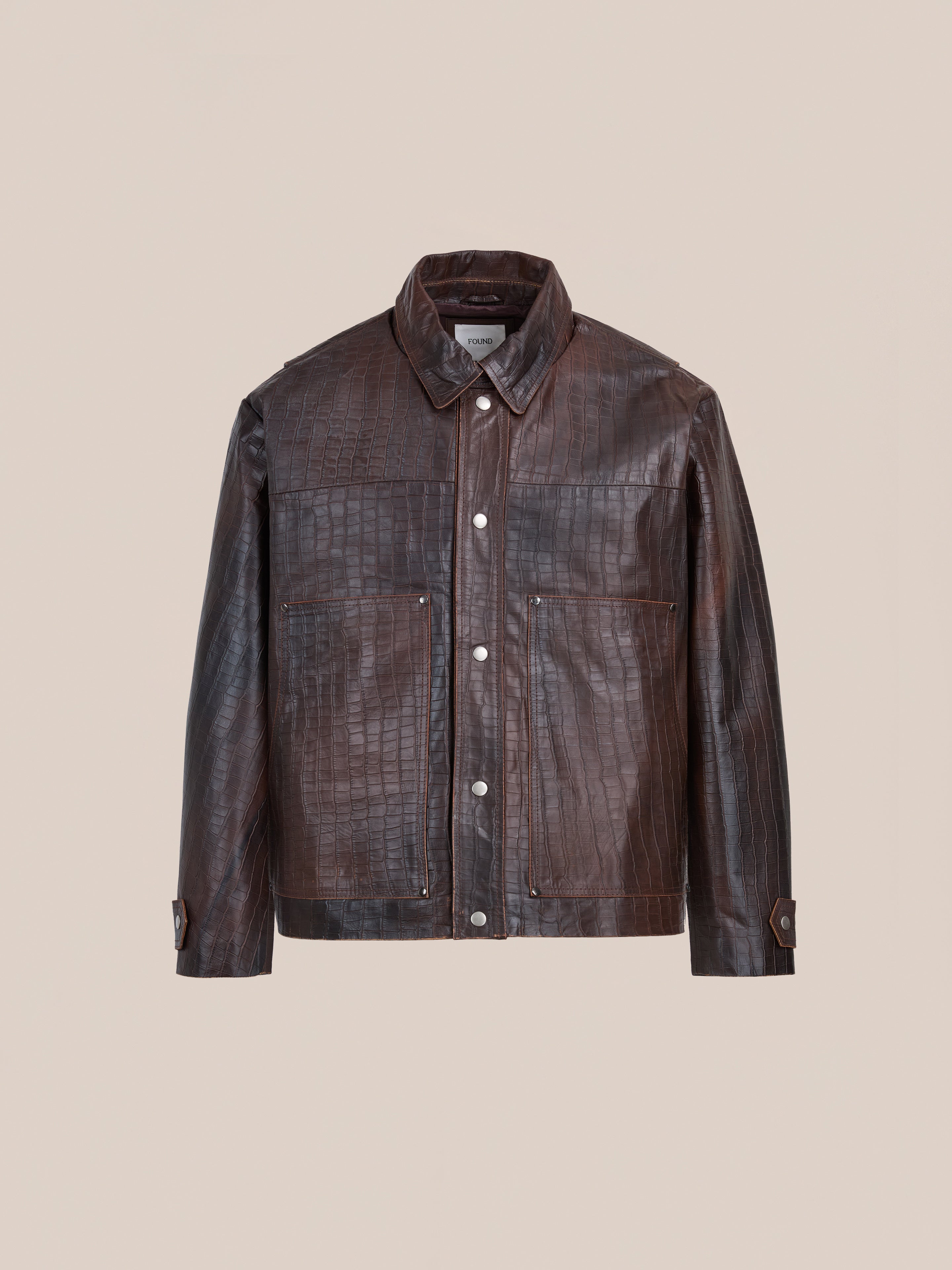 The FOUND Distressed Croc Embossed Leather Work Jacket features a collar, front snap buttons, two chest pockets, and buttoned cuffs. This unisex design combines classic workwear style with bold croc texture against a plain background.
