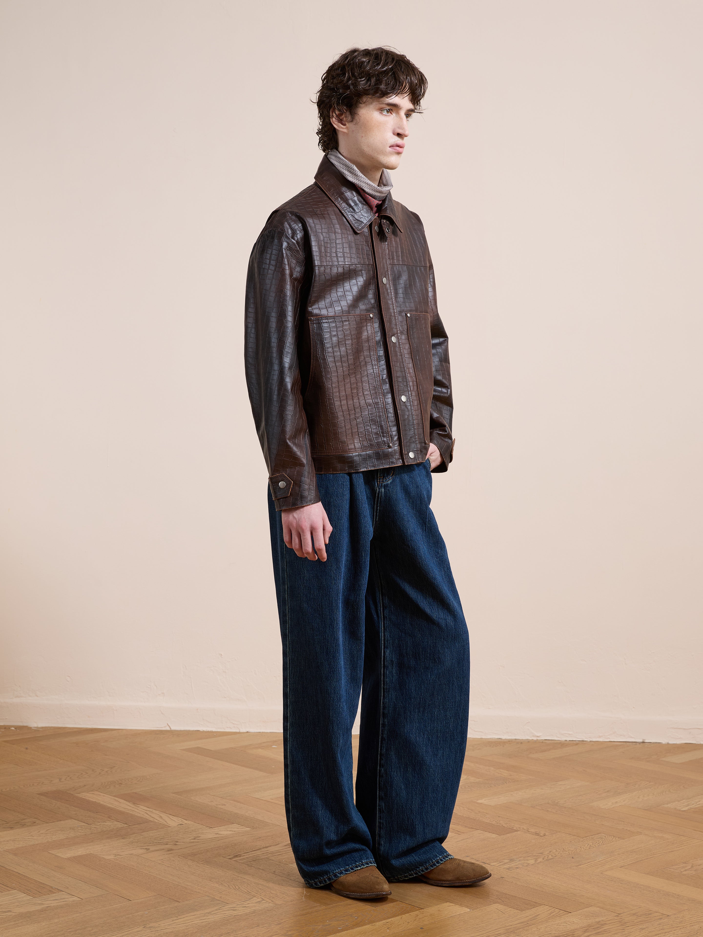 Someone stands indoors against a plain beige wall and wooden floor, wearing the FOUND Distressed Croc Embossed Leather Work Jacket, blue wide-leg jeans, and brown shoes.