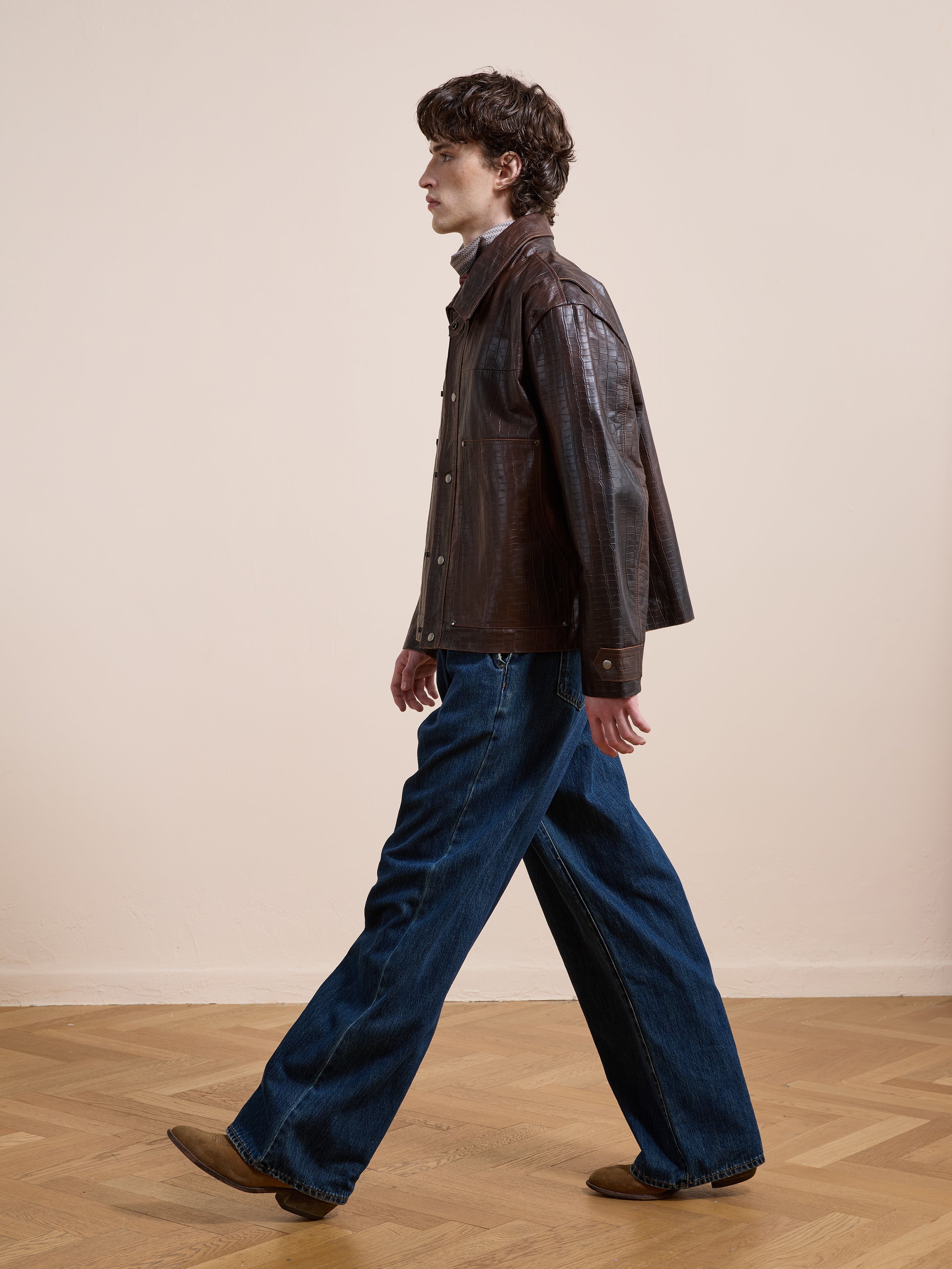 A person in a FOUND Distressed Croc Embossed Leather Work Jacket, wide-leg jeans, and brown shoes walks indoors on a wooden floor with a plain beige wall behind them.