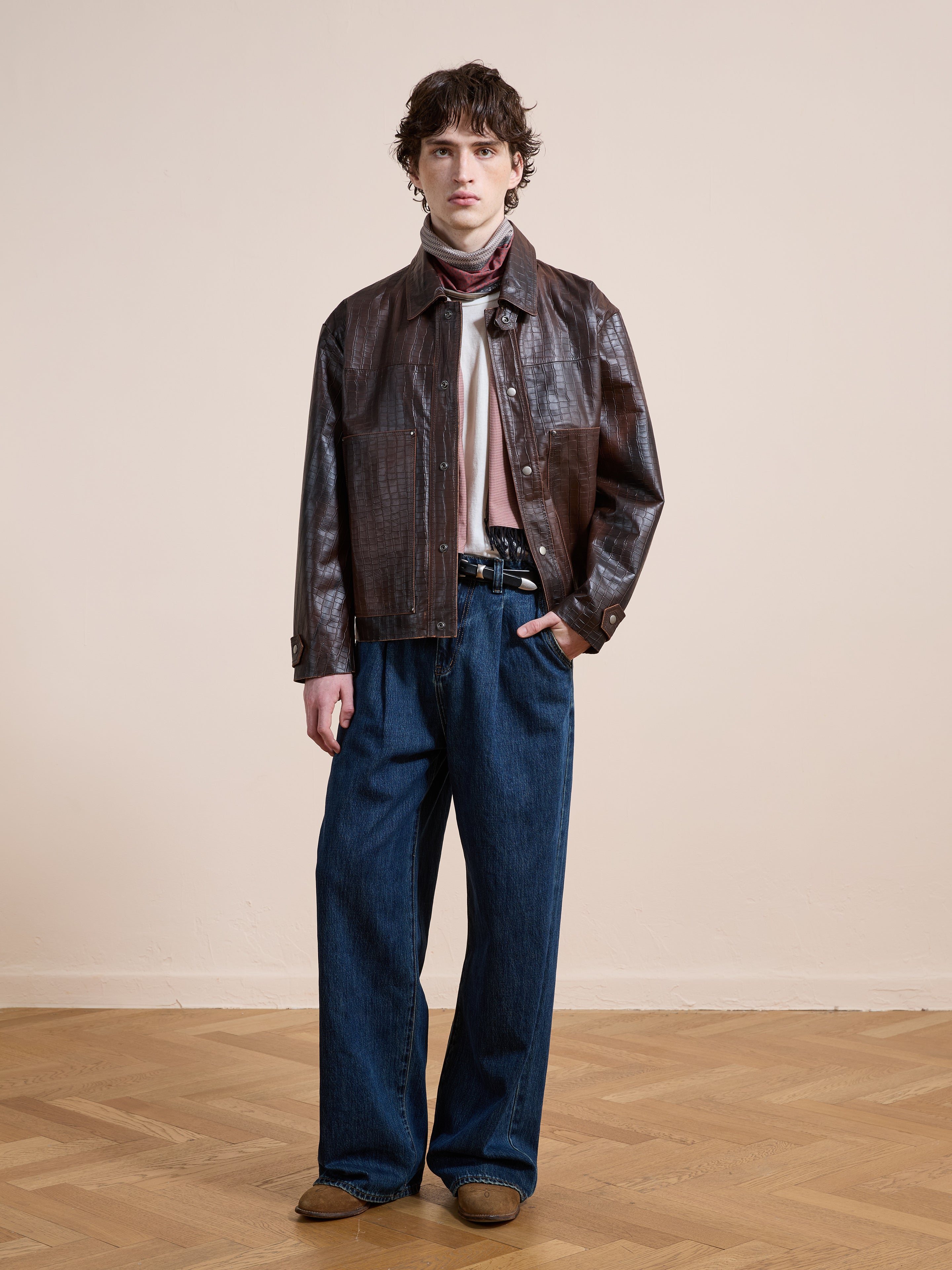 Indoors, a person wears the FOUND Distressed Croc Embossed Leather Work Jacket with wide-leg blue jeans, a light shirt, scarf, and brown shoes on a wooden floor against a plain beige wall.