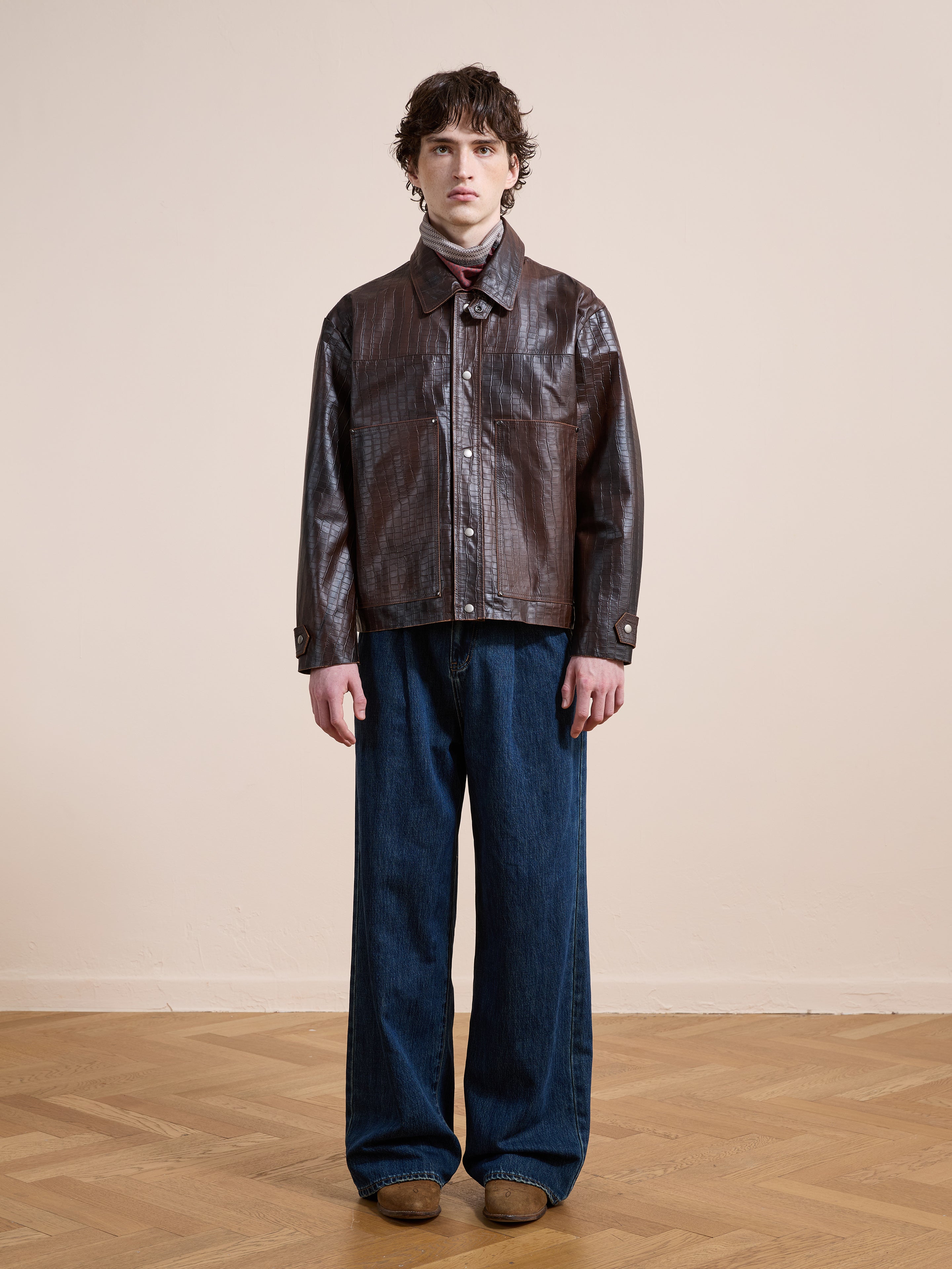 Indoors on a wooden floor with a beige wall, a person wears the FOUND Distressed Croc Embossed Leather Work Jacket, styled with a scarf, wide-leg blue jeans, and brown shoes.