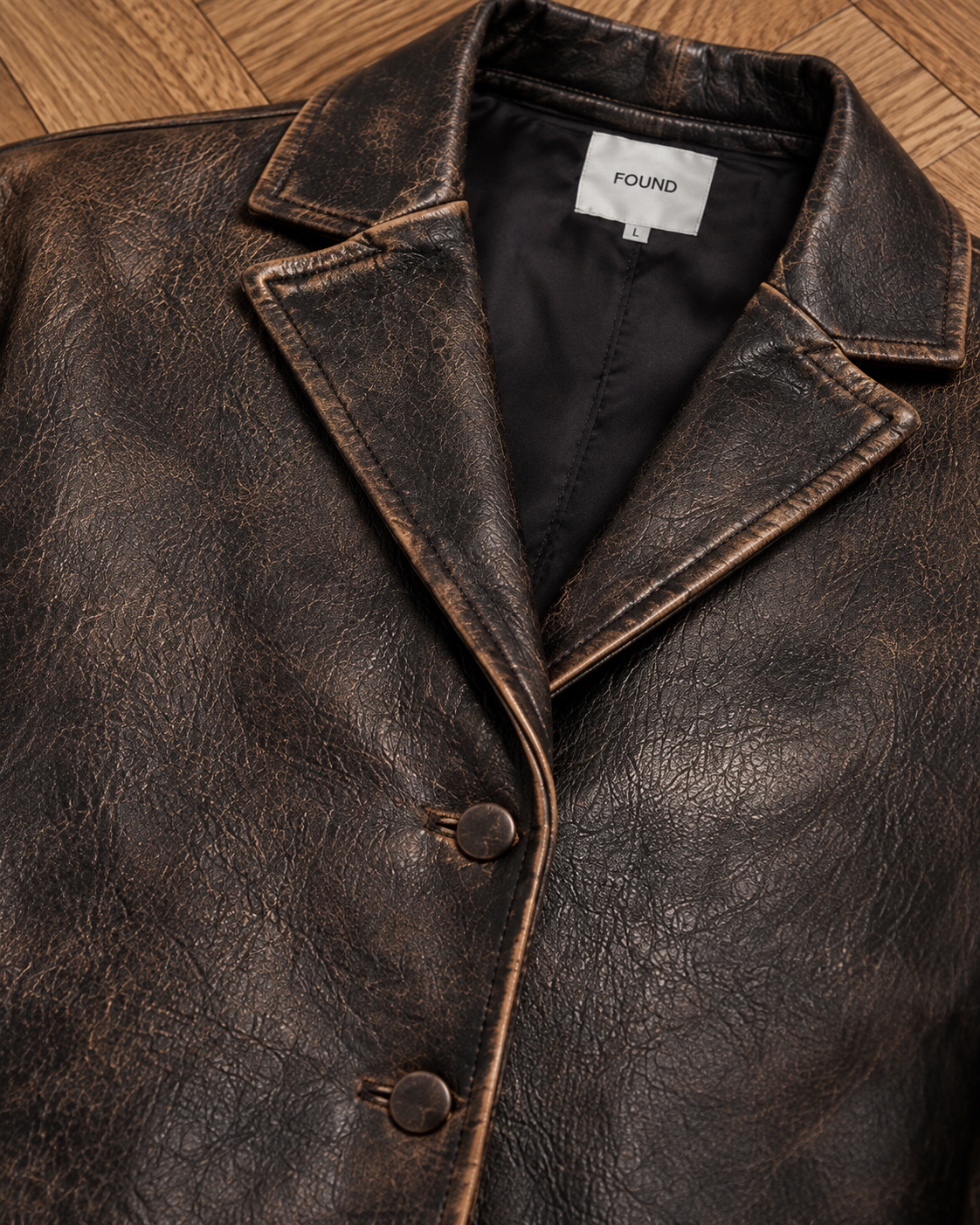 Close-up of a Distressed Leather Blazer by FOUND, size large, with two buttons and brand label visible on the inside collar, laid flat on a wooden floor.