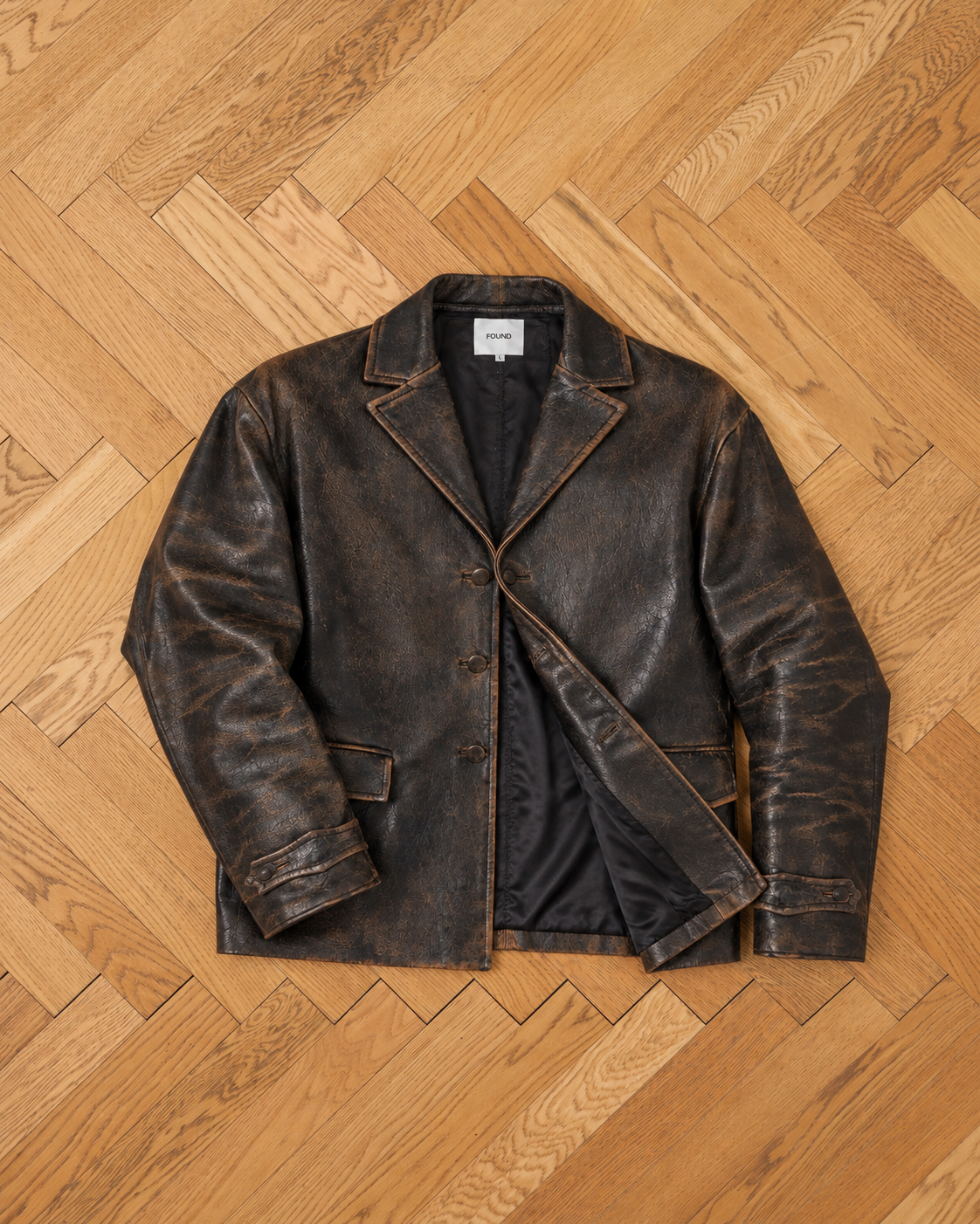 The FOUND Distressed Leather Blazer, featuring a button front, notched lapels, and front pockets, is laid flat on a light wooden herringbone floor.