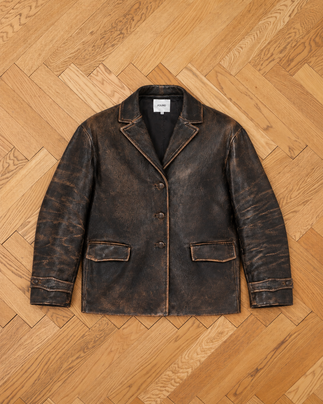 The FOUND Distressed Leather Blazer features three front buttons and two side pockets, displayed on a light wood herringbone floor.
