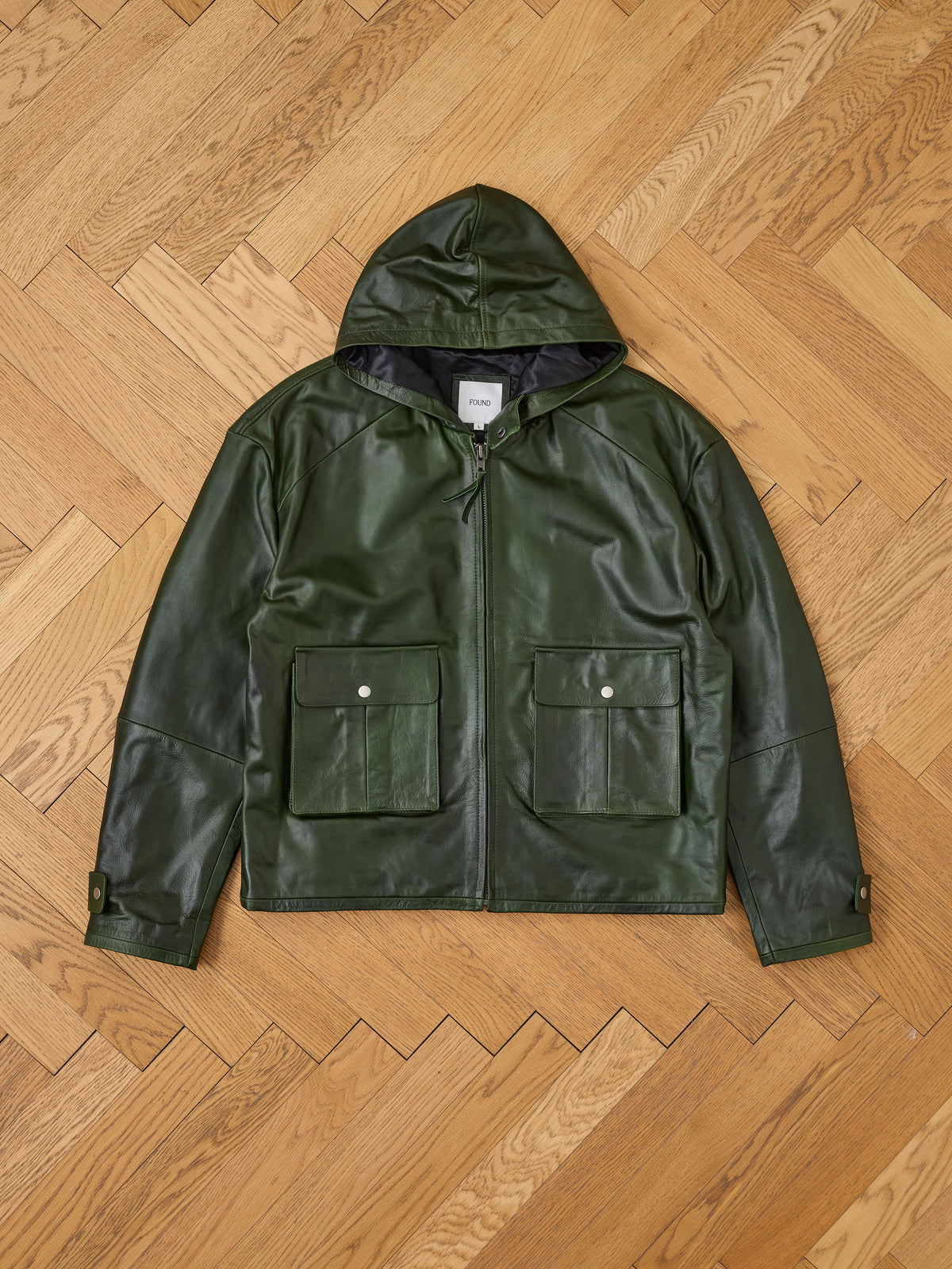 The FOUND Distressed Leather Pocket Hoodie, featuring a dark green leather finish, two front pockets, and snap-button cuffs, is displayed on a wooden herringbone floor.