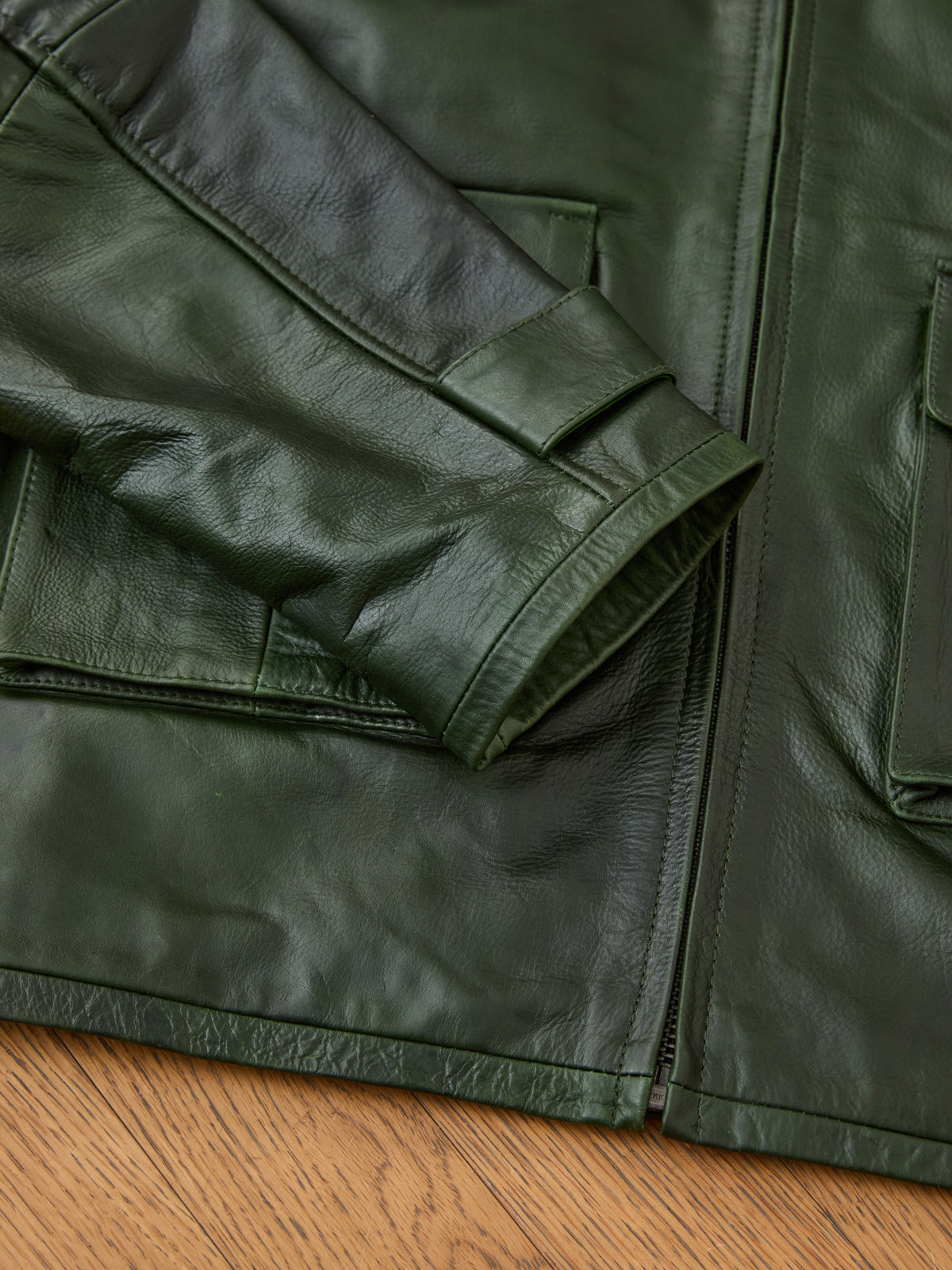 Close-up of the FOUND Distressed Leather Pocket Hoodie in dark green, highlighting the sleeve and pocket details, laid flat on a wooden surface.
