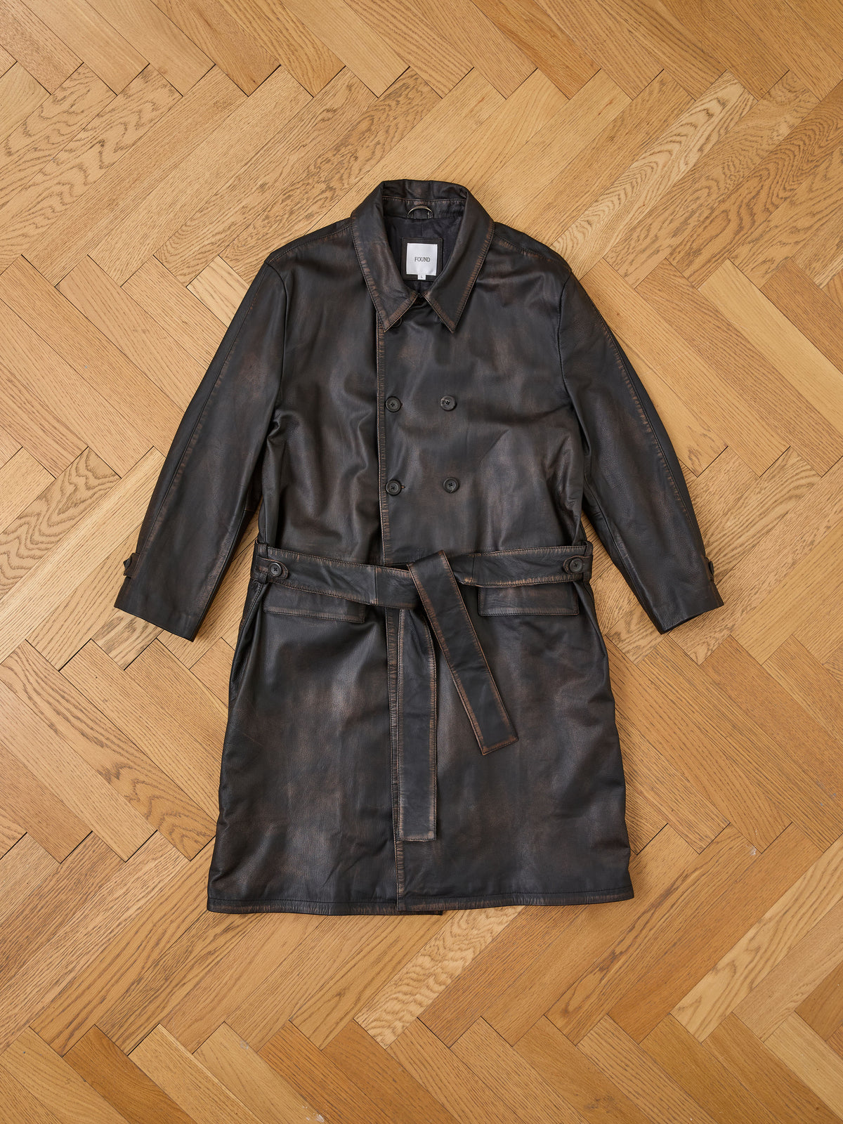 The FOUND Belted Leather Trench Coat in dark brown, ethically crafted, is shown laid flat on a light wood herringbone floor.
