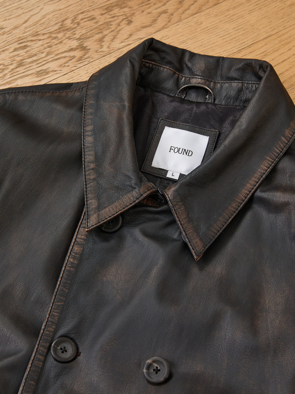 Belted Leather Trench Coat by FOUND with double-breasted buttons and collar, laid flat on a light wood surface.