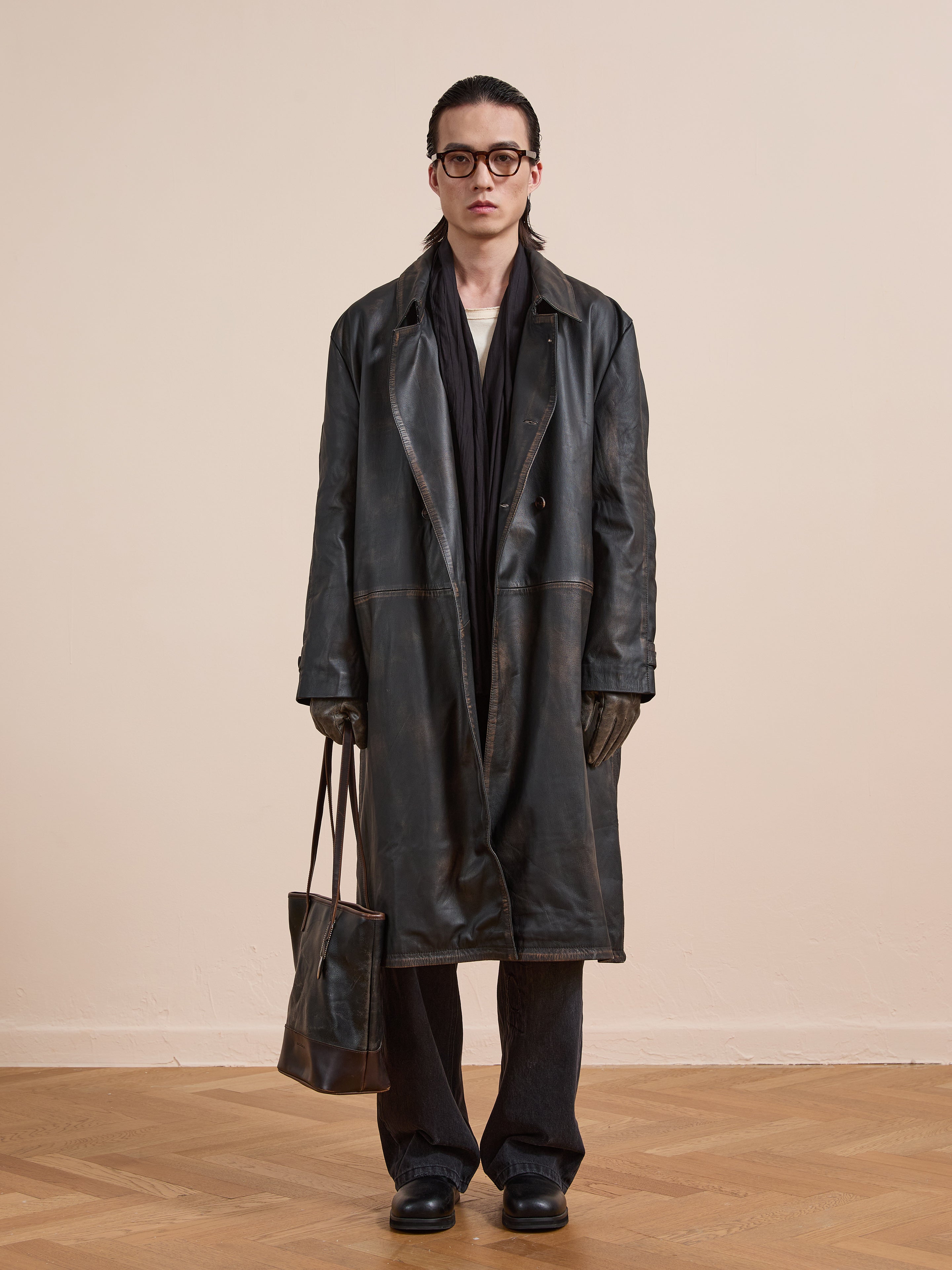 A person stands indoors against a plain wall, wearing the FOUND Distressed Leather Trench Coat, gloves, glasses, and holding a brown bag.