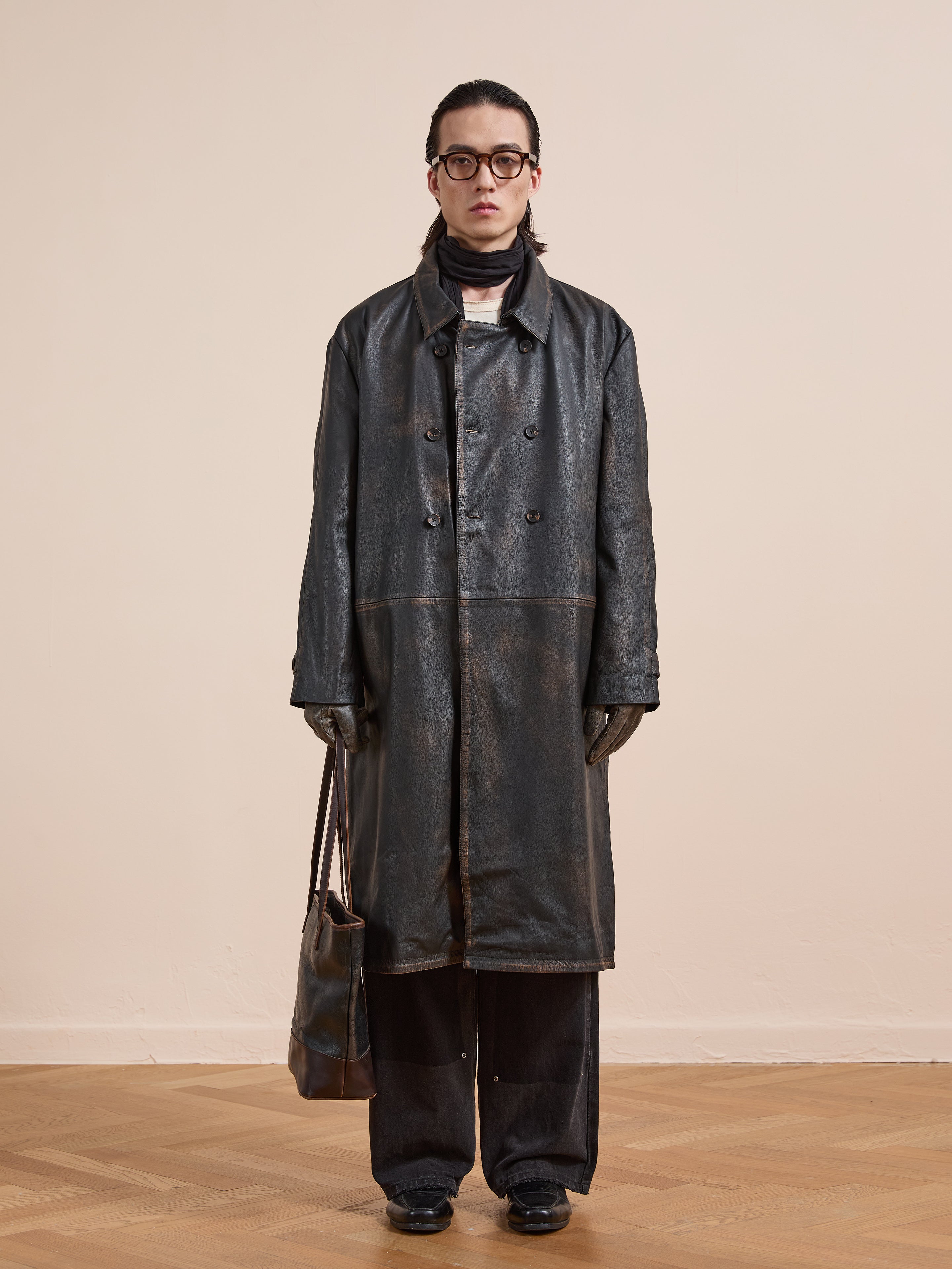 Indoors against a beige wall and wooden floor, a person wears FOUND's Distressed Leather Trench Coat with black pants, glasses, scarf, gloves, and holds a brown bag—the coat lending an effortlessly stylish edge to the outfit.
