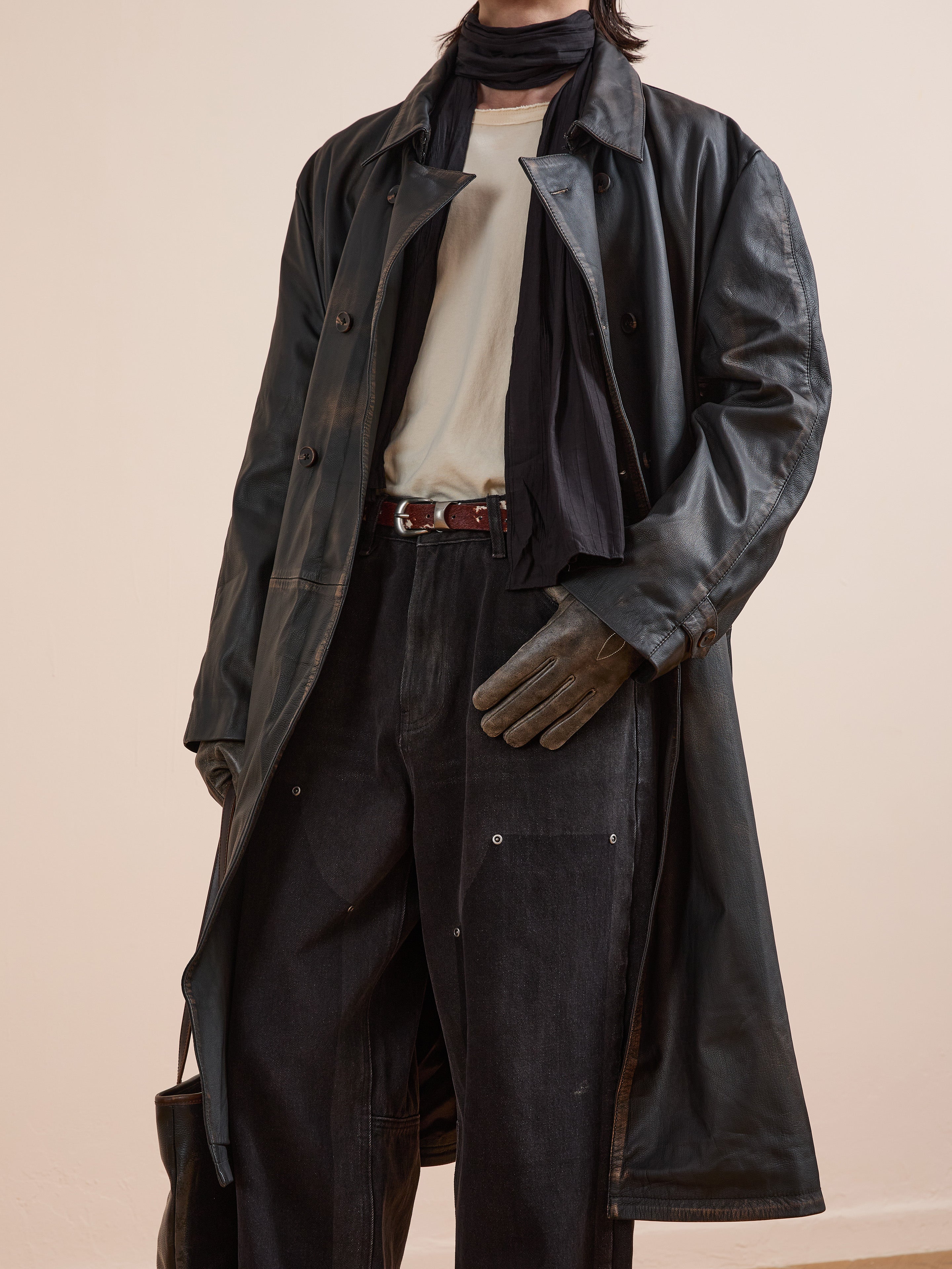 A person wears FOUND’s Distressed Leather Trench Coat with a white shirt, black scarf, dark pants, and brown gloves, holding a bag in one hand.