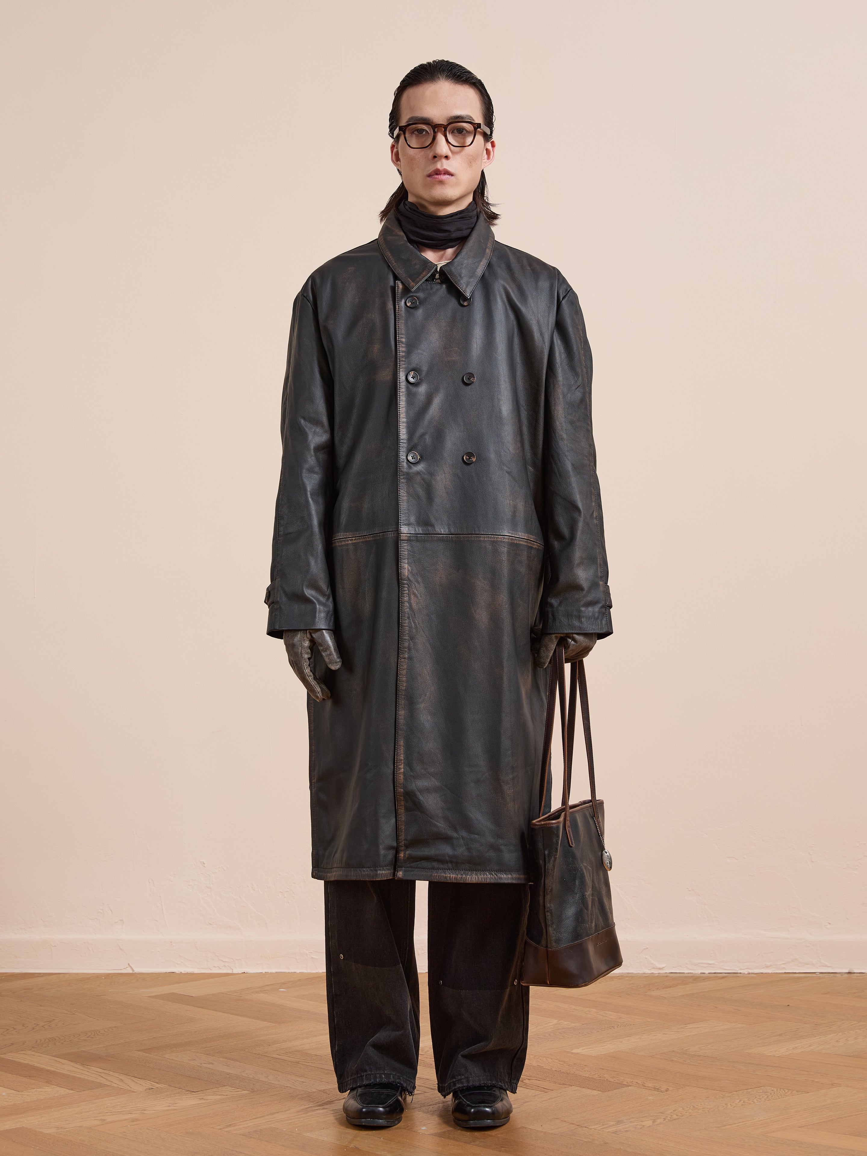 A person stands indoors facing the camera against a plain background, wearing the FOUND Distressed Leather Trench Coat, black pants, gloves, glasses, and holding a brown tote bag.
