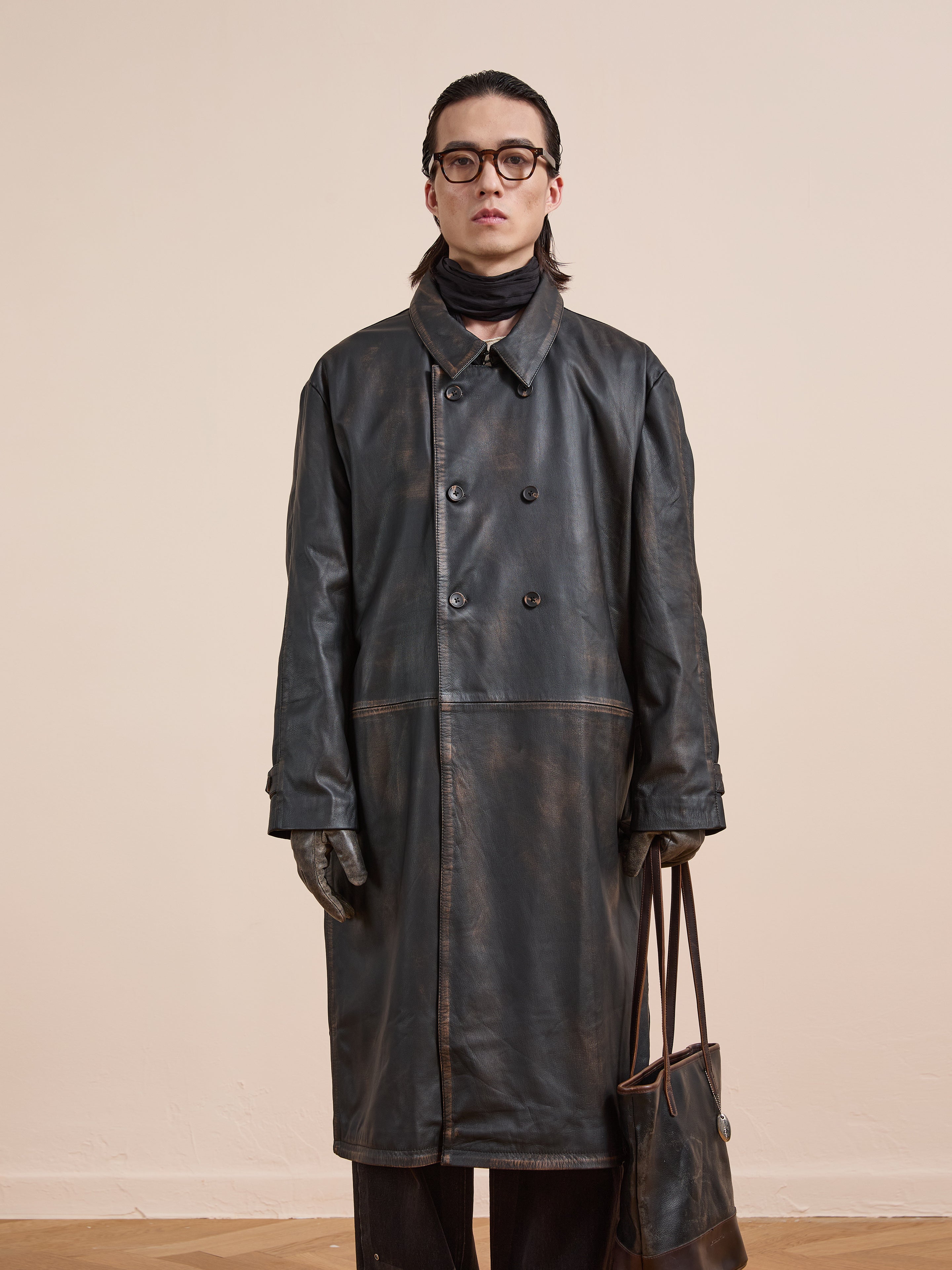 A person wears the FOUND Distressed Leather Trench Coat, gloves, glasses, and a scarf while standing against a plain background and holding a brown leather tote bag.
