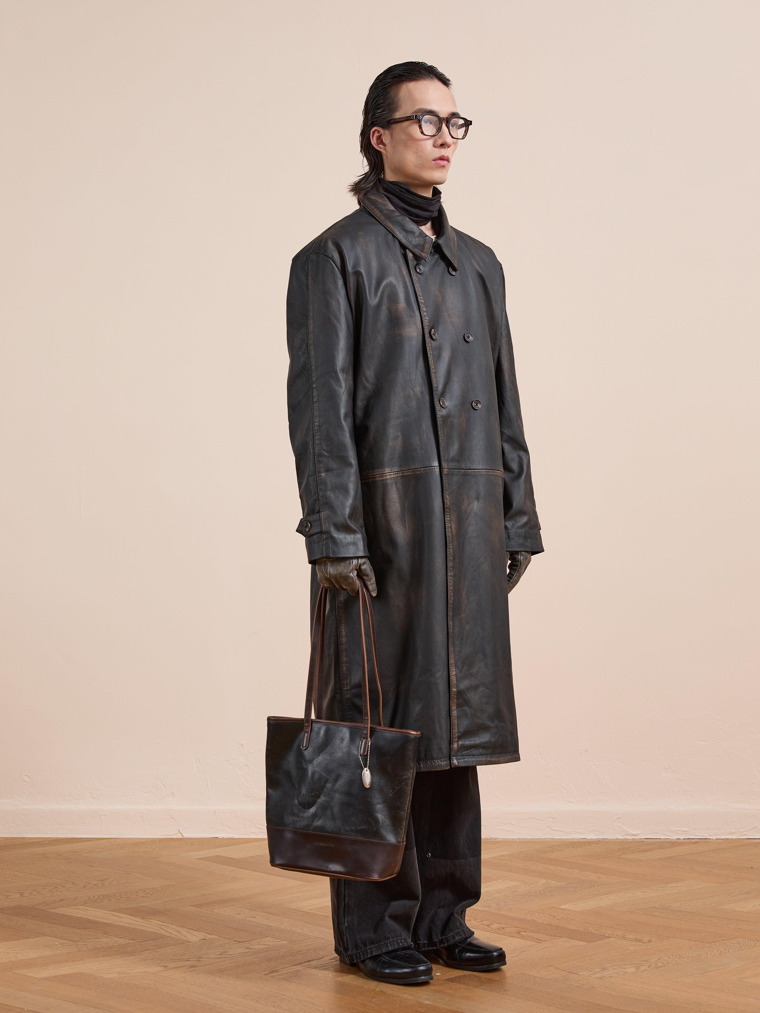 A person wears the FOUND Distressed Leather Trench Coat, black pants, gloves, and glasses, holding a brown leather tote bag while standing against a beige wall on a wooden floor.