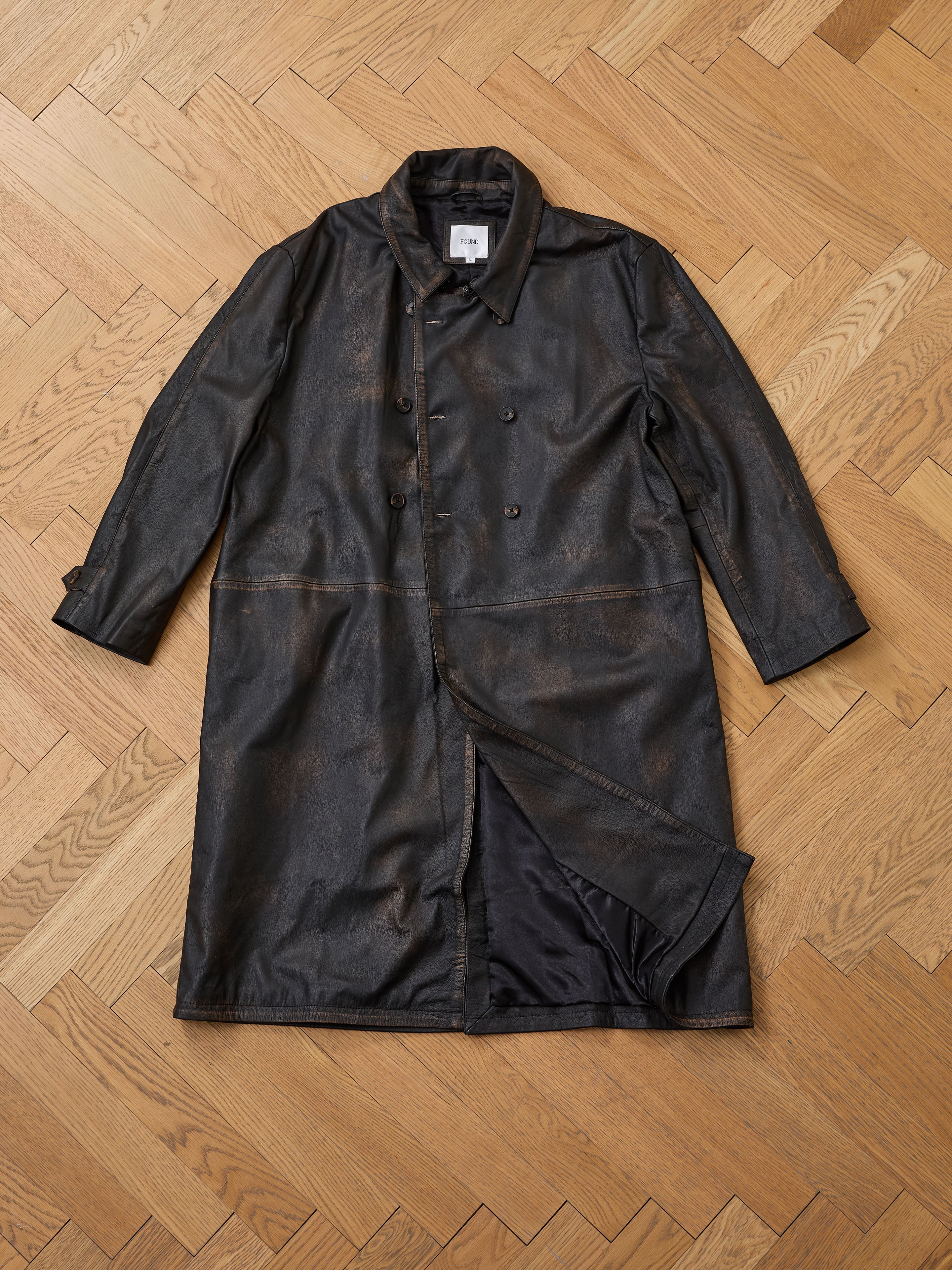 The FOUND Distressed Leather Trench Coat, featuring a double-breasted front and wide collar, is laid flat on a light wooden herringbone floor, highlighting its hand-distressed black leather finish.