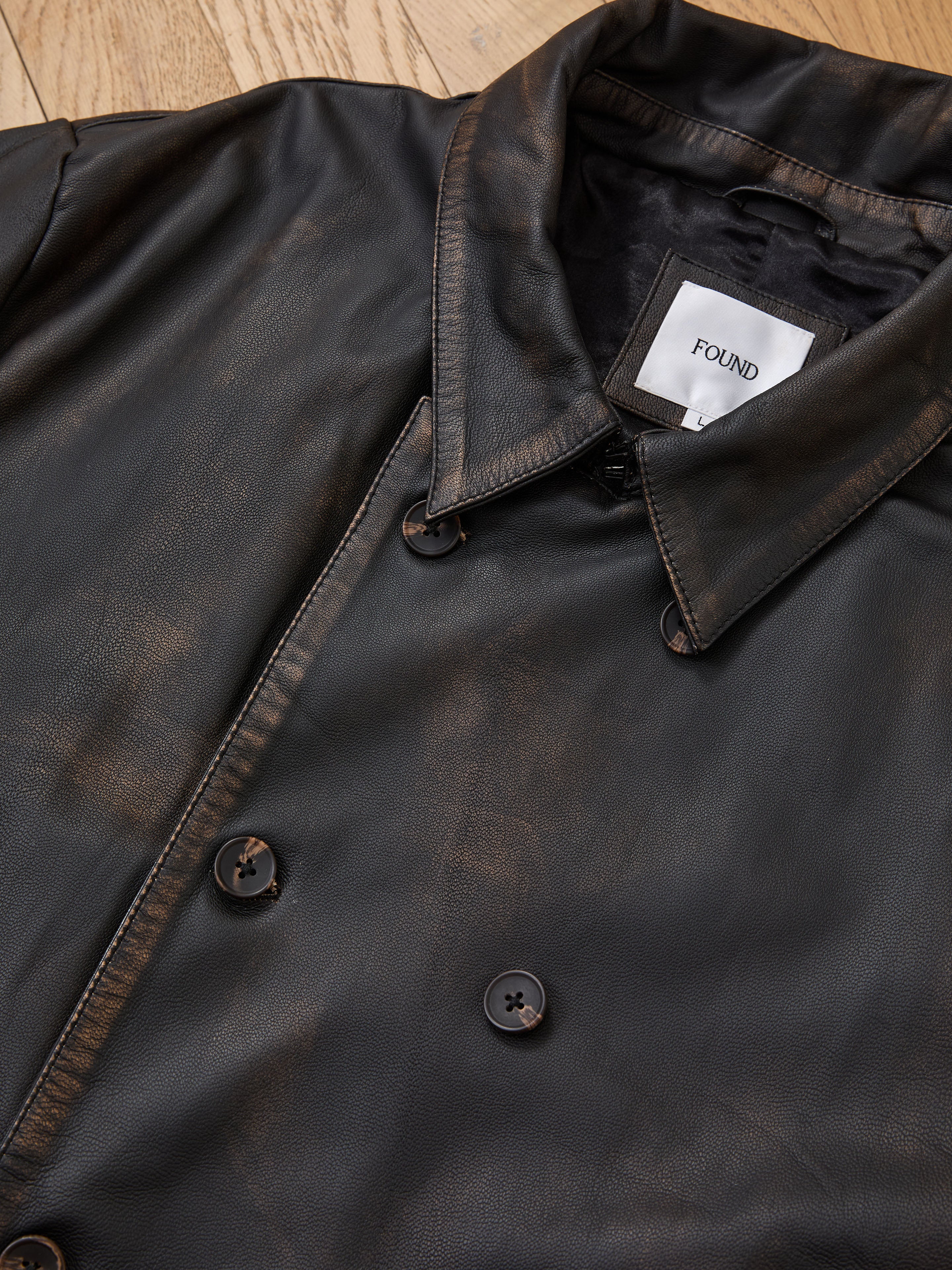 A close-up of the FOUND Distressed Leather Trench Coat, black and laid flat on a wooden surface.