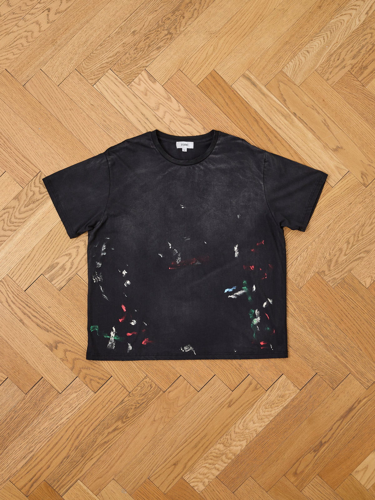 The Distressed Paint Tee by FOUND is a black, short-sleeve unisex relaxed fit T-shirt featuring subtle paint stains in a variety of colors. Laid flat on a wooden herringbone-patterned floor, this tee exudes a casual, artistic vibe, making it an ideal choice for everyday wear.