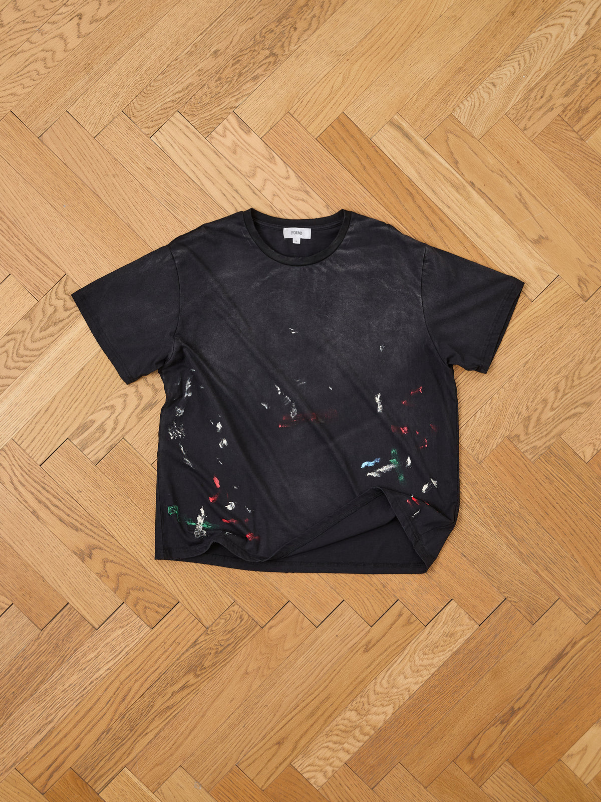 A FOUND Distressed Paint Tee, featuring black fabric with scattered paint splatters, is laid flat on a wooden herringbone floor.