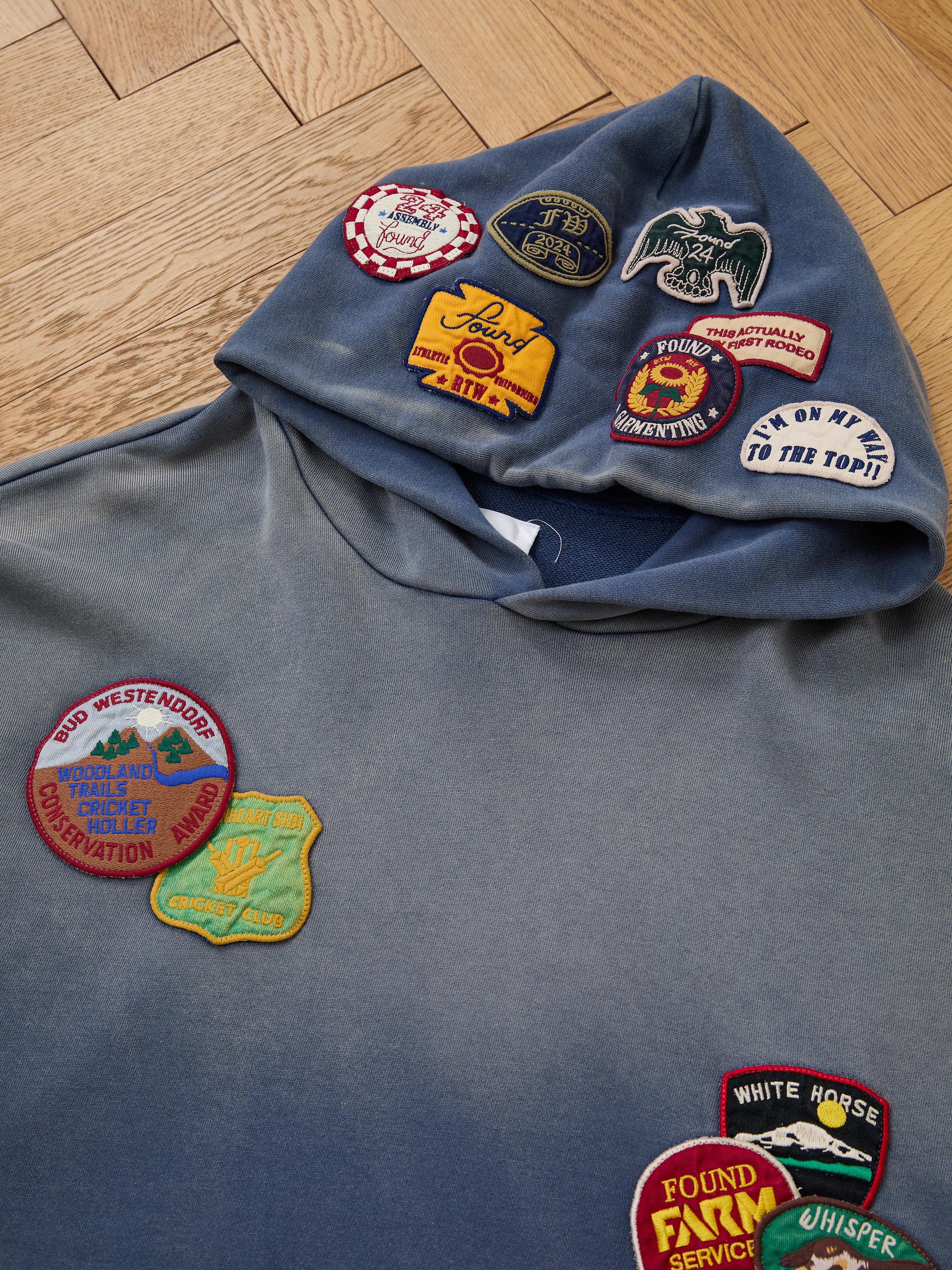 The FOUND Distressed Patch Hoodie is a blue French Terry cotton hoodie featuring colorful embroidered patches on the hood and chest, shown laid flat on a wooden floor.