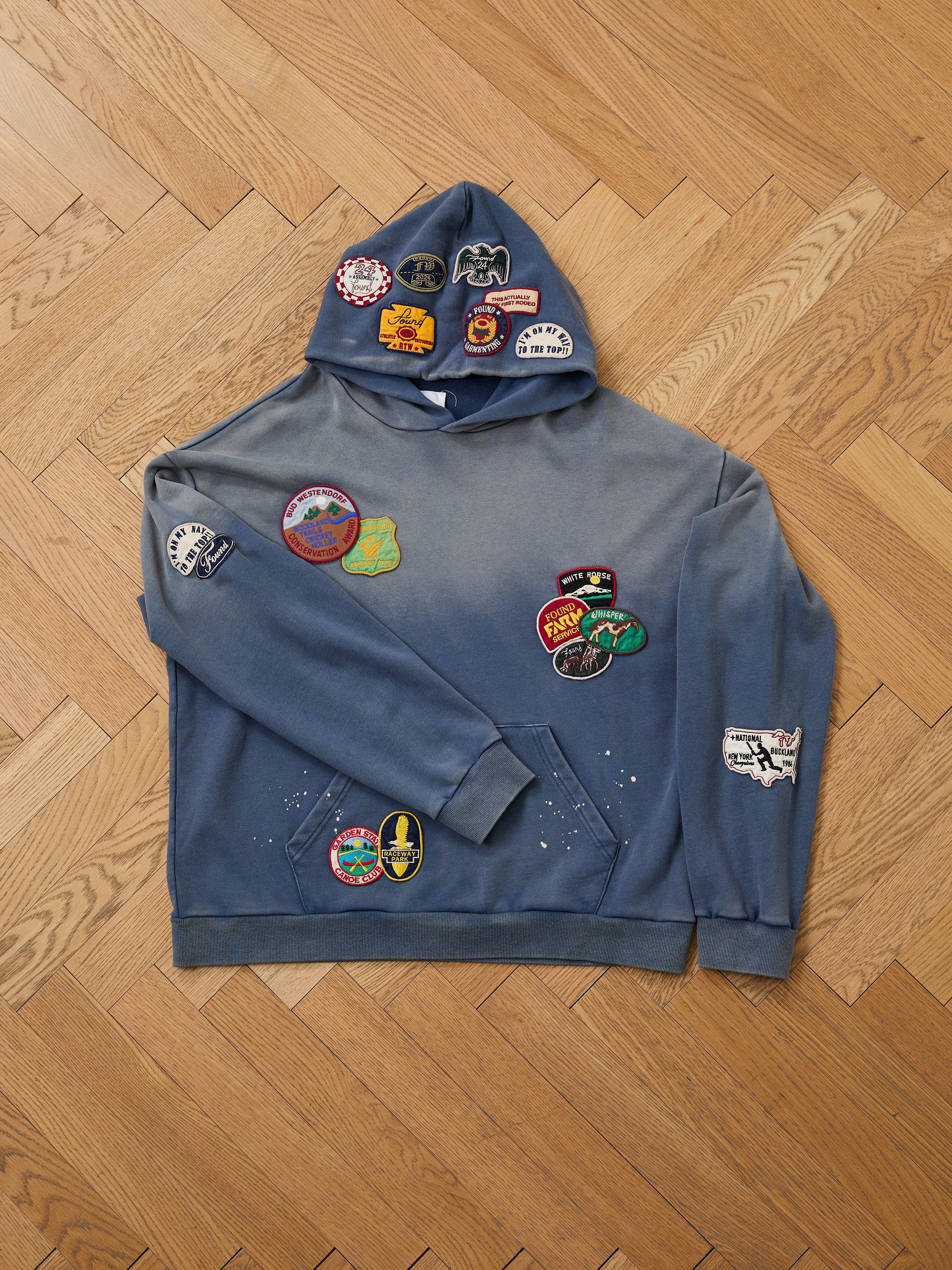 The FOUND Distressed Patch Hoodie in blue, made of French Terry cotton, features colorful embroidered patches on the front, hood, and left sleeve, displayed flat on a wooden herringbone floor.